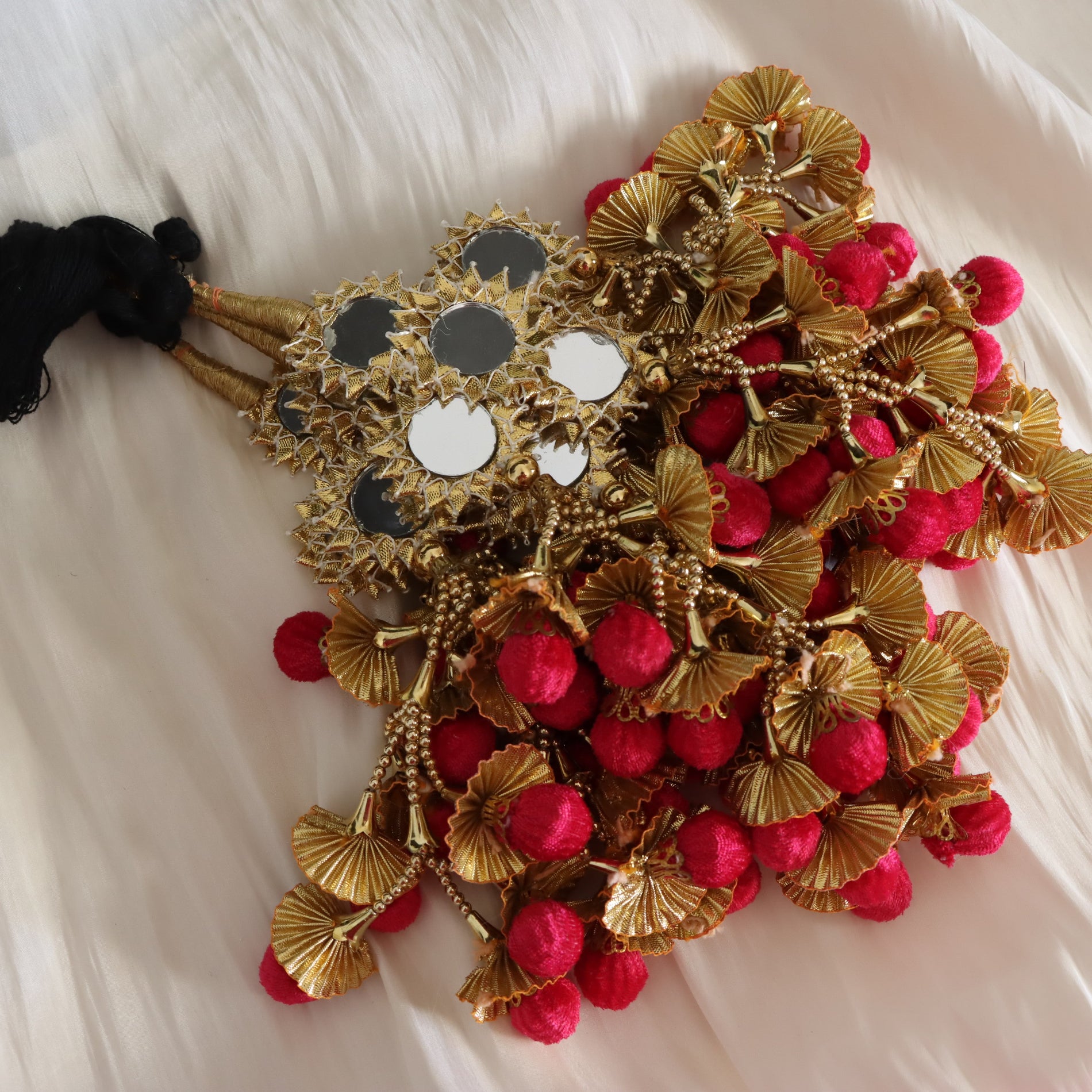 A decorative bridal hairpiece or headpiece laid flat on a white fabric background. The piece features gold metallic fan-shaped ornaments, small gold beads, clusters of bright pink pom-pom berries, and several round black-and-white mirrored discs attached to a black fabric tie at the left.