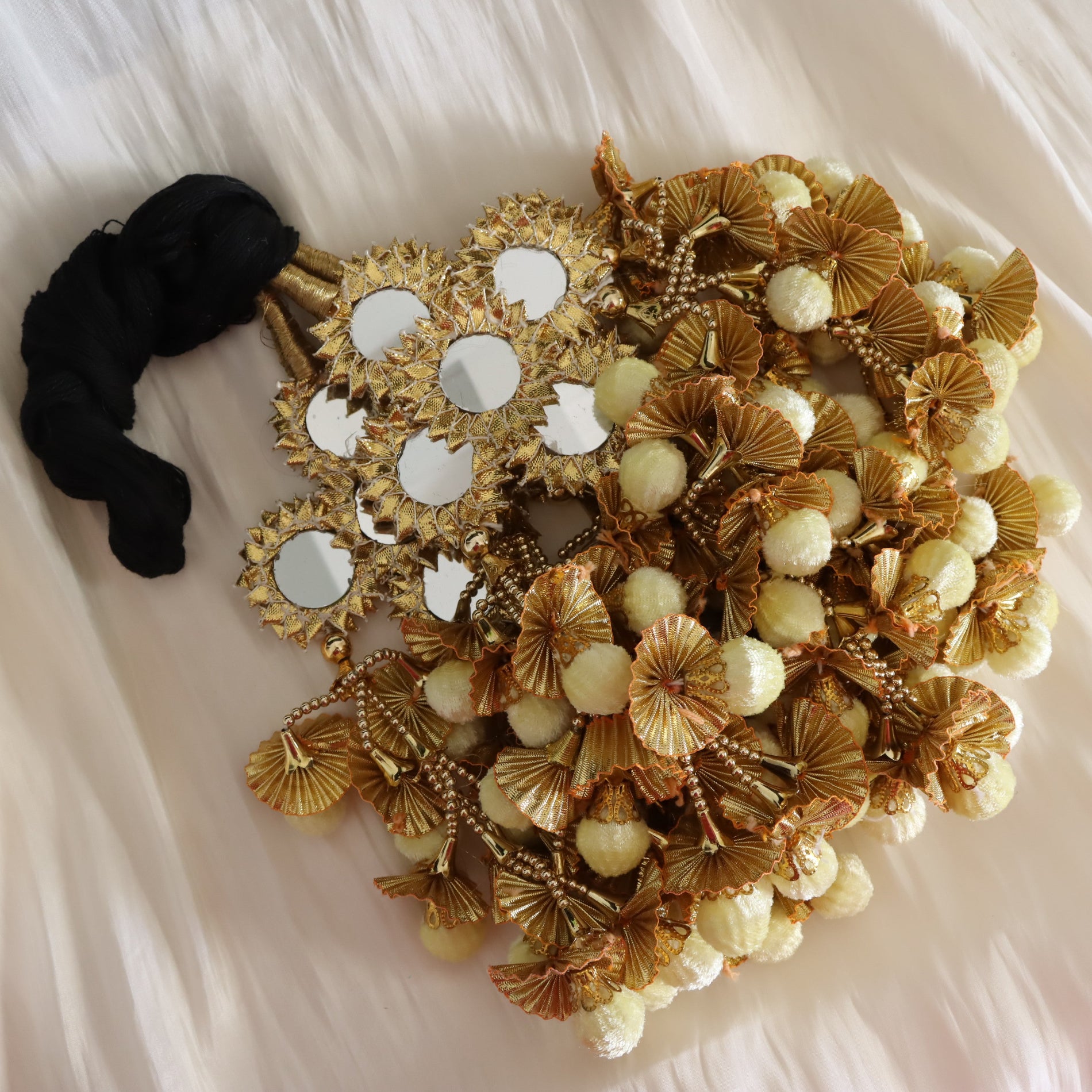 A decorative bridal bouquet head made from gold artificial palm leaves, cream pom-poms, beaded strands, and several small round mirrors with gold frames, arranged on a white fabric background next to a black fabric bow.
