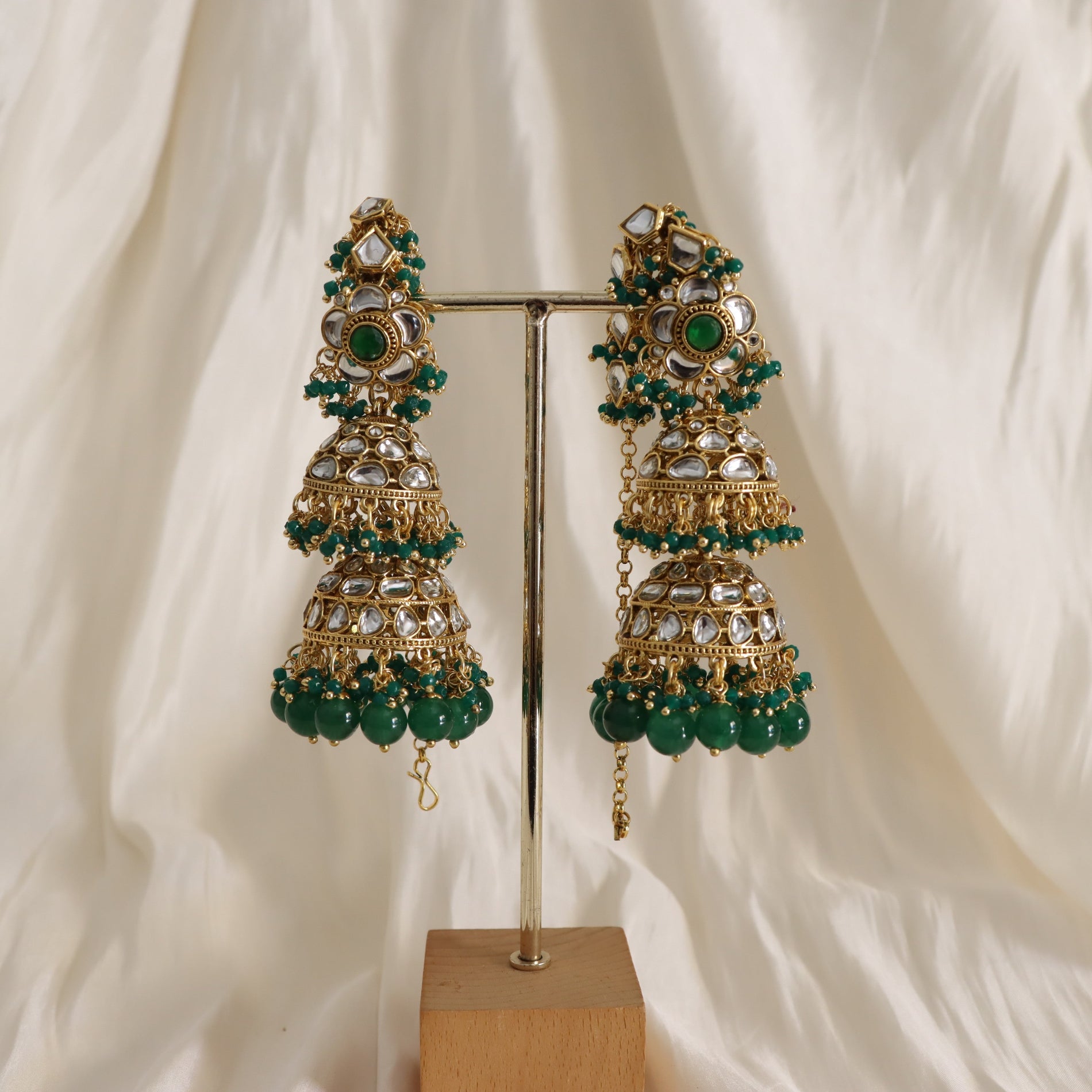 Ornate earrings with green beads on a stand against a white curtain background