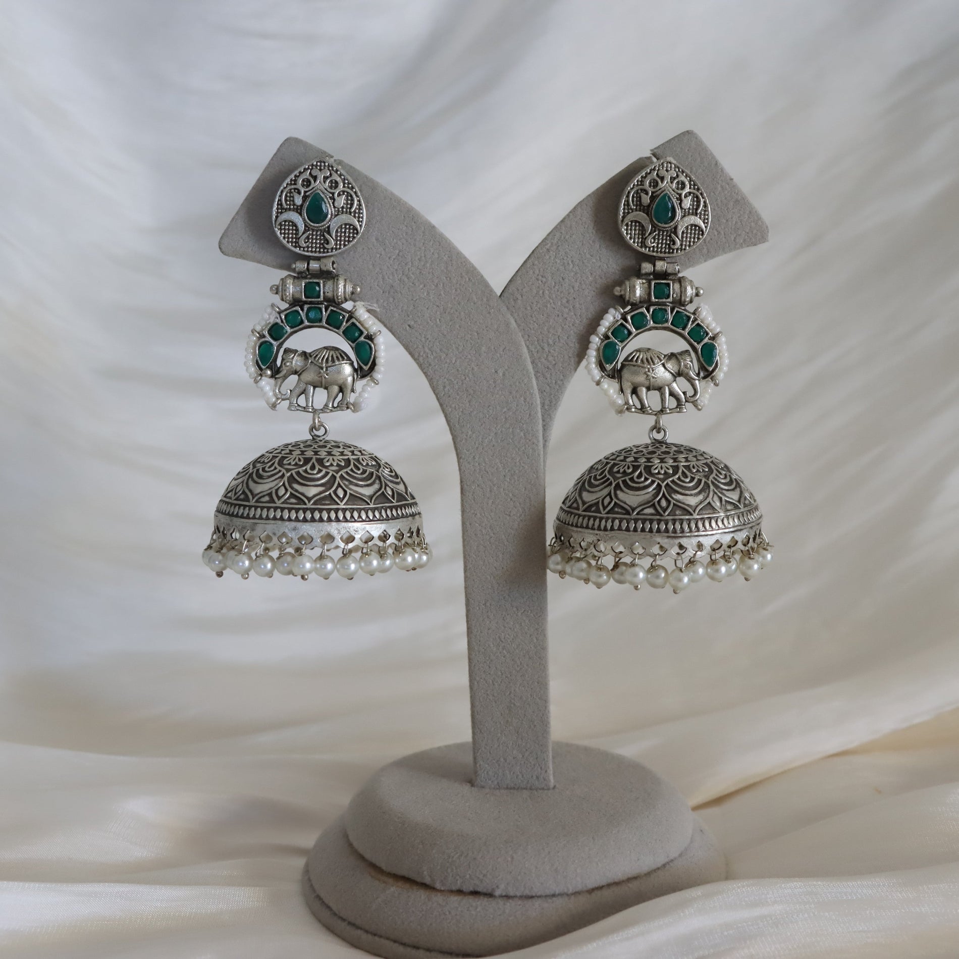 Silver-toned Indian jhumka earrings with green stone accents and white pearl fringe, displayed on a gray velvet jewelry stand against a draped white fabric background.
