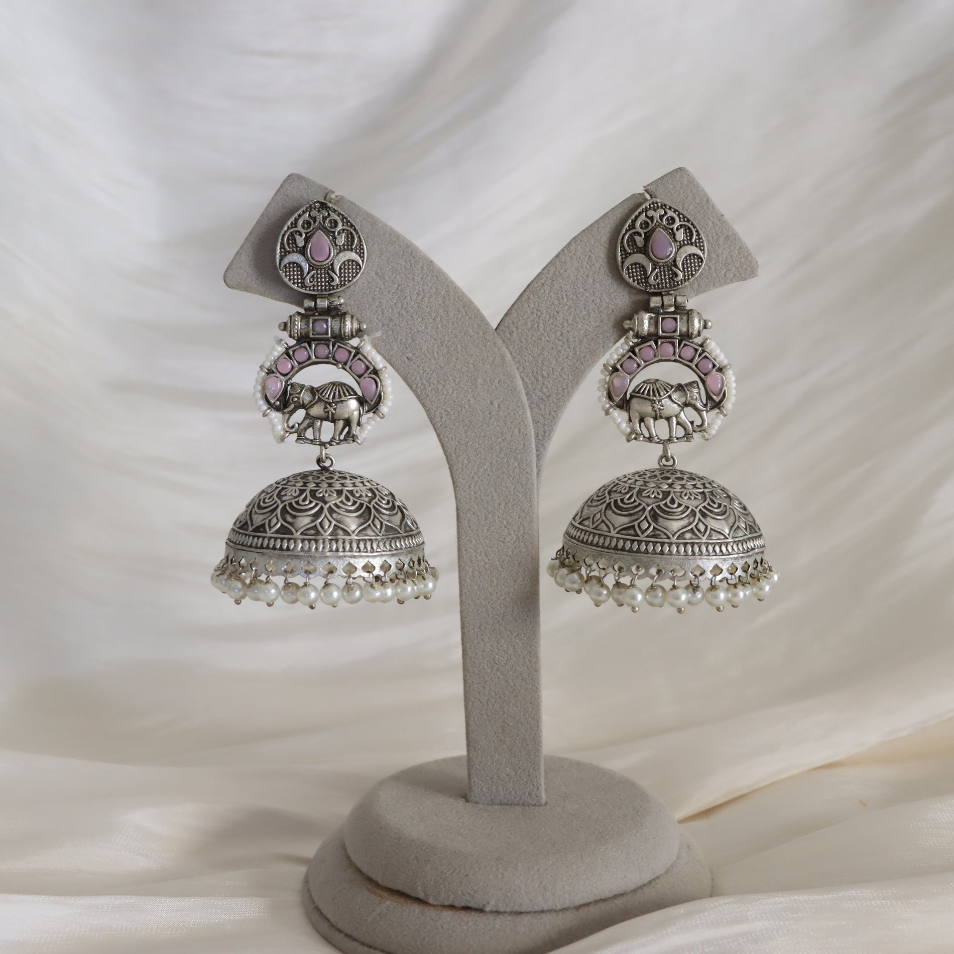A pair of ornate silver-toned Indian jhumka earrings with pink enamel, elephant motifs, and white pearl bead borders, displayed on a gray velvet jewelry stand against a draped white fabric background.