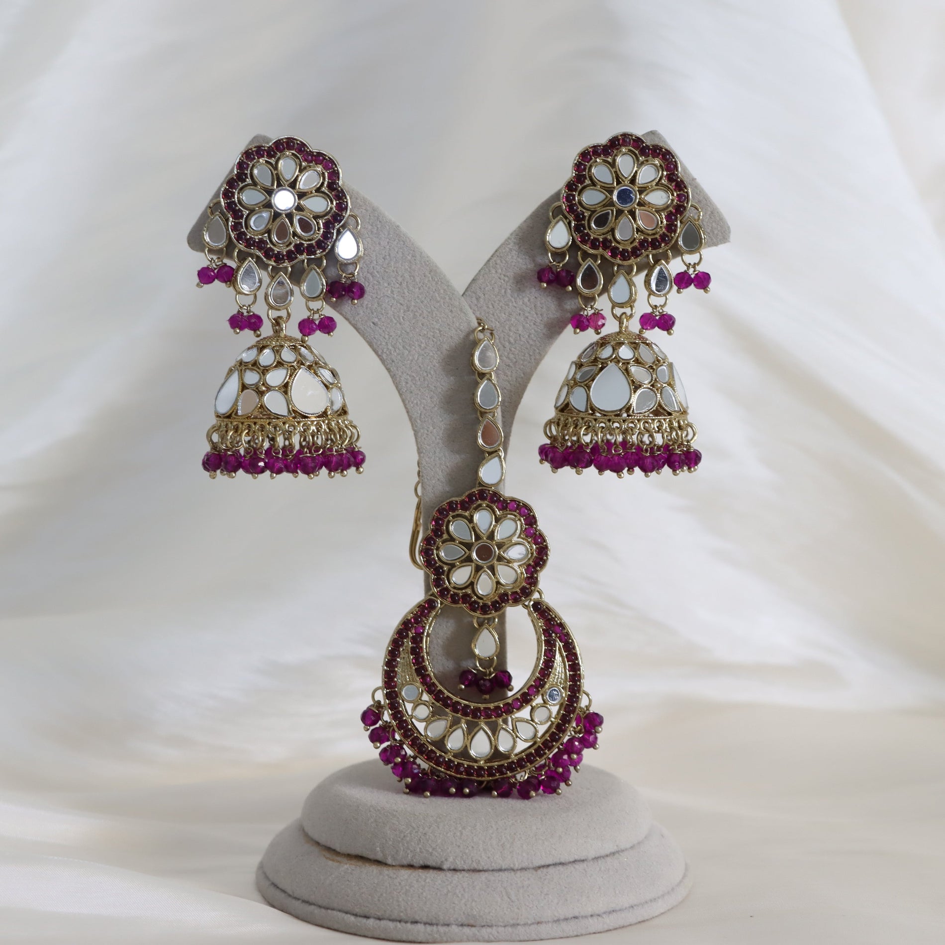 A pair of ornate Indian jhumka earrings displayed on a light gray jewelry stand against a draped white fabric background. The earrings feature gold-plated metal work, white stone inlay, and deep purple bead accents.