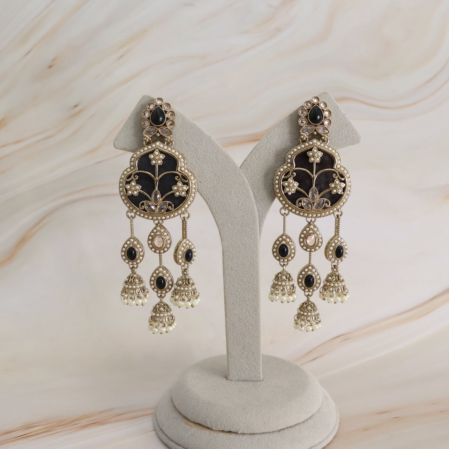 A pair of ornate gold-toned dangle earrings with black enamel inlay and small white bead accents, displayed on a light gray jewelry stand against a soft cream marble background.