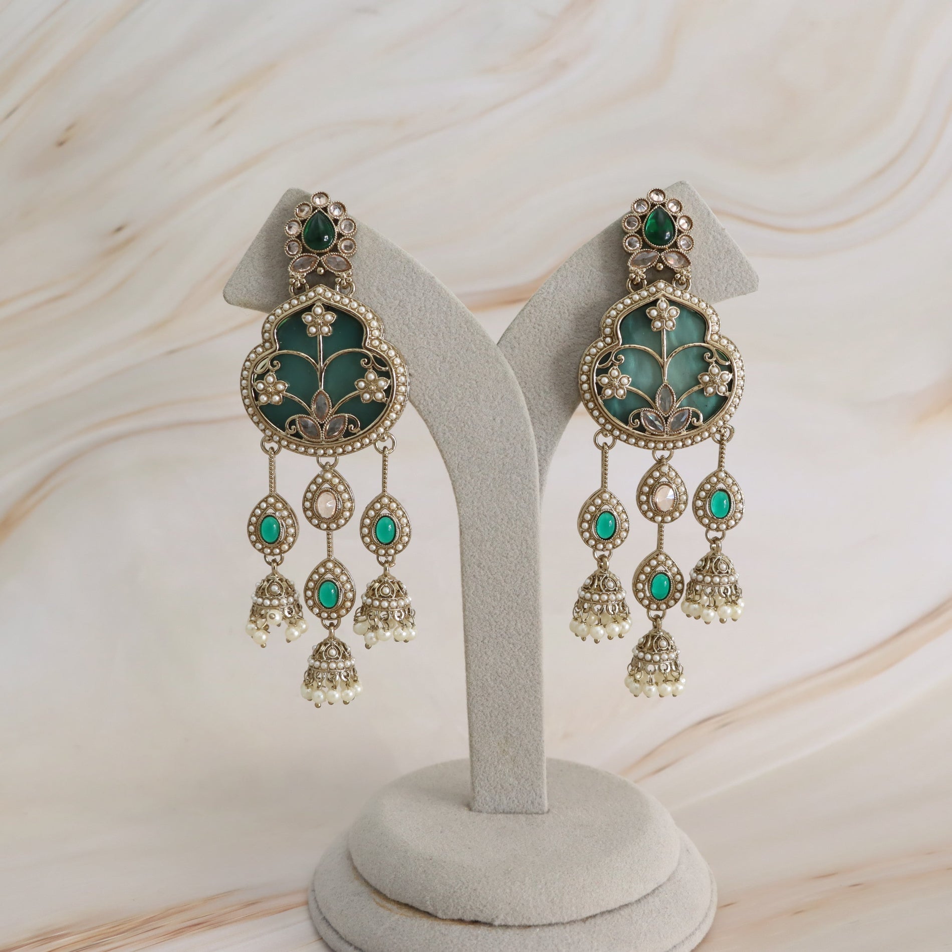 A pair of ornate gold-toned dangle earrings with green enamel panels and small pearl-like accents, displayed on a light gray jewelry stand against a softly marbled cream background.