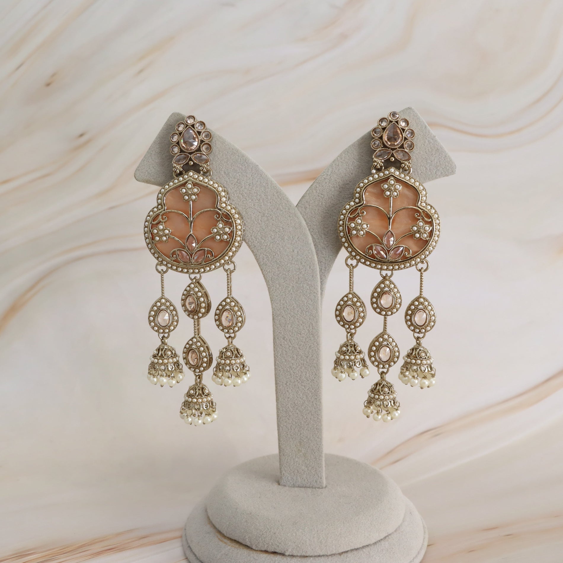 A pair of ornate gold-toned Indian jhumka earrings with peach-pink stone inlays and white pearl-like bead drops, displayed on a light gray jewelry stand against a cream marble background.