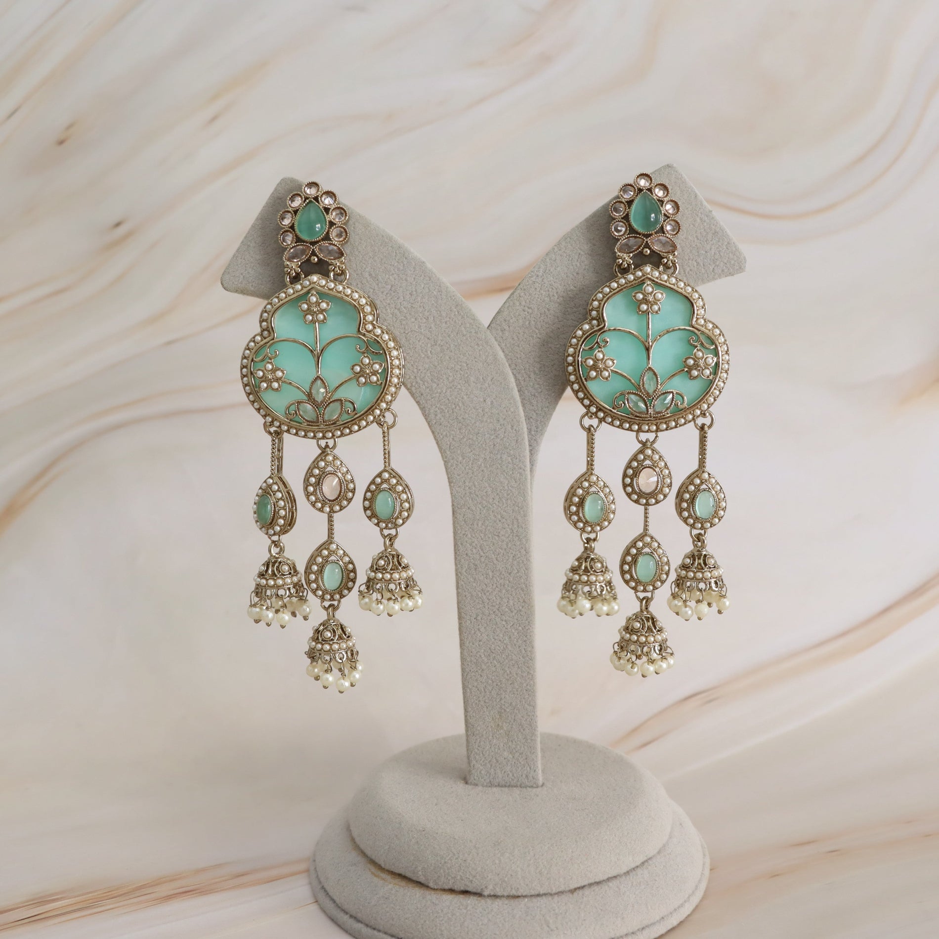 A pair of ornate dangle earrings displayed on a light gray jewelry stand. The earrings feature mint green enamel inlays, gold-toned metalwork, and small white bead details, set against a light beige marble-patterned background.