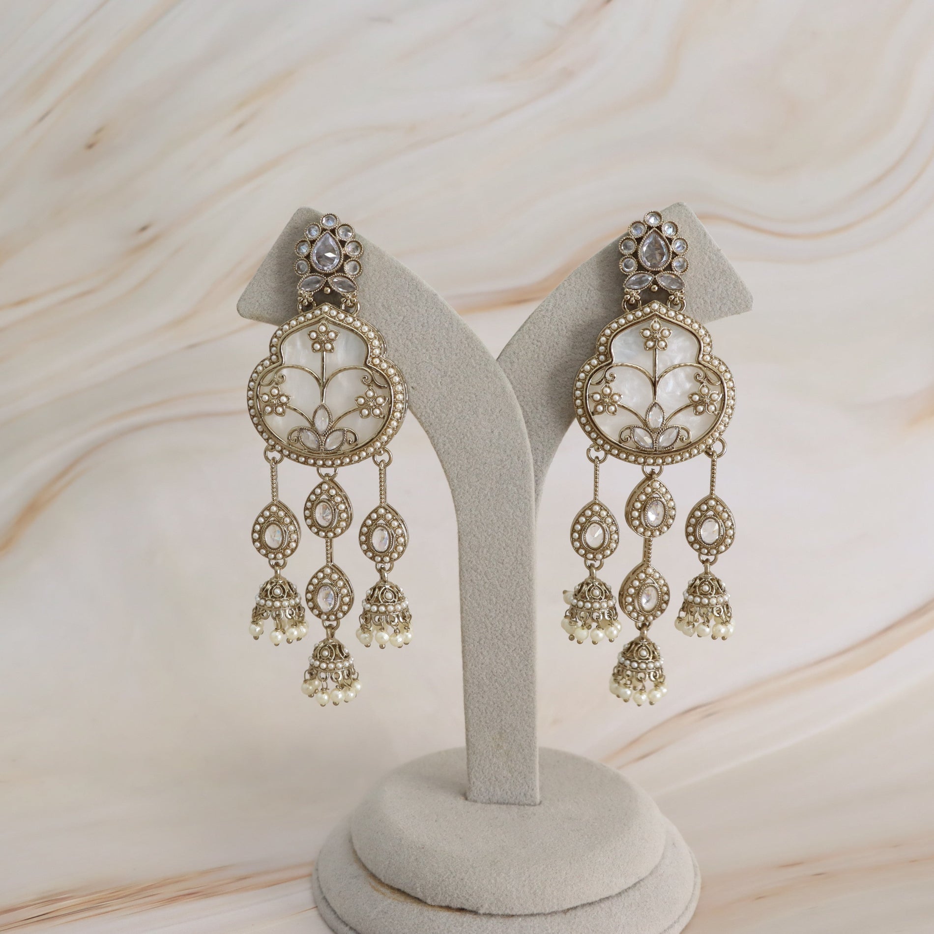 A pair of ornate gold-toned dangle earrings with white pearl-like accents, displayed on a light gray jewelry stand against a soft beige marble-patterned background.