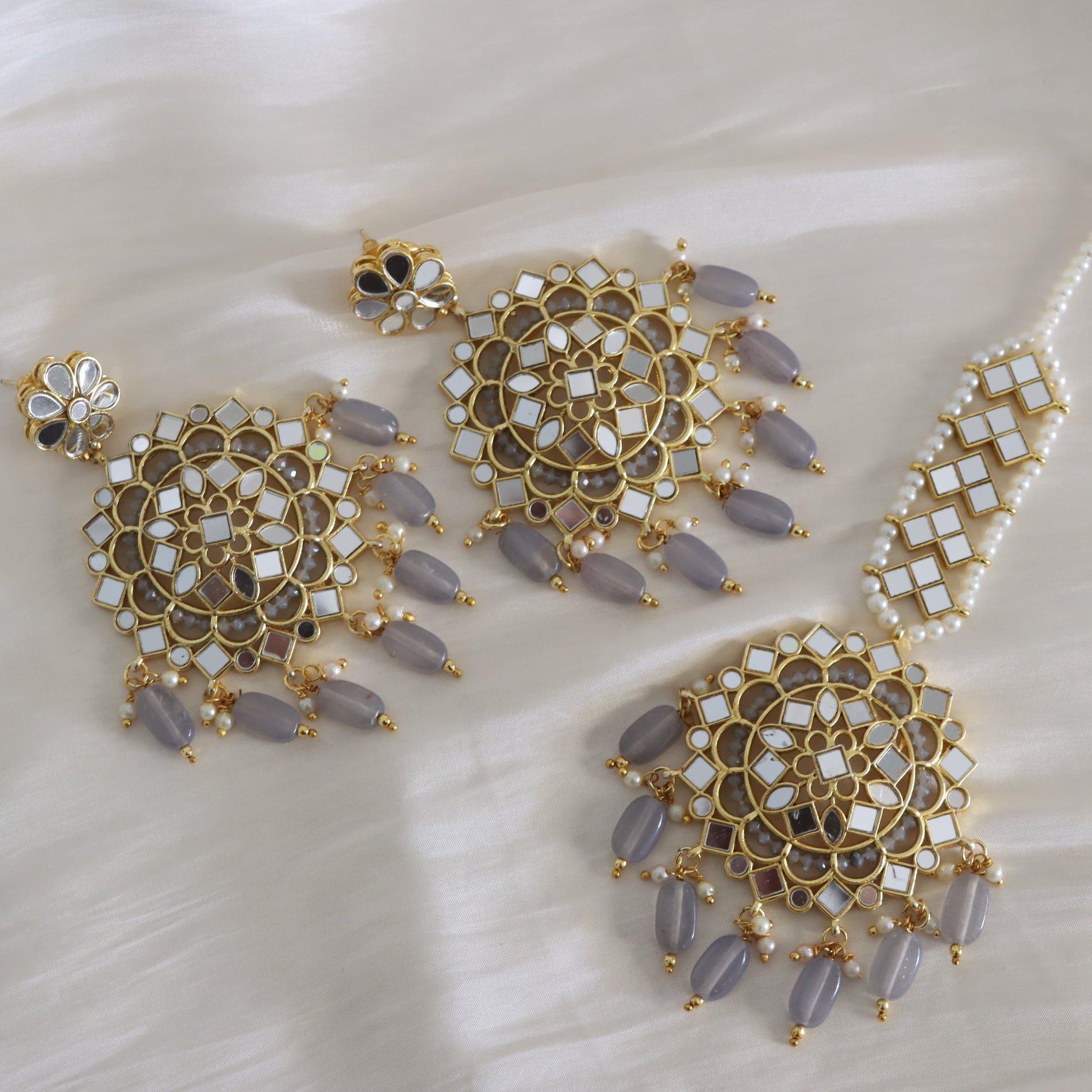 Gold-toned traditional Indian jewelry set featuring a large choker necklace, matching earrings, and a bracelet, decorated with white inlay stones and lavender-gray bead tassels, displayed on a white satin fabric.