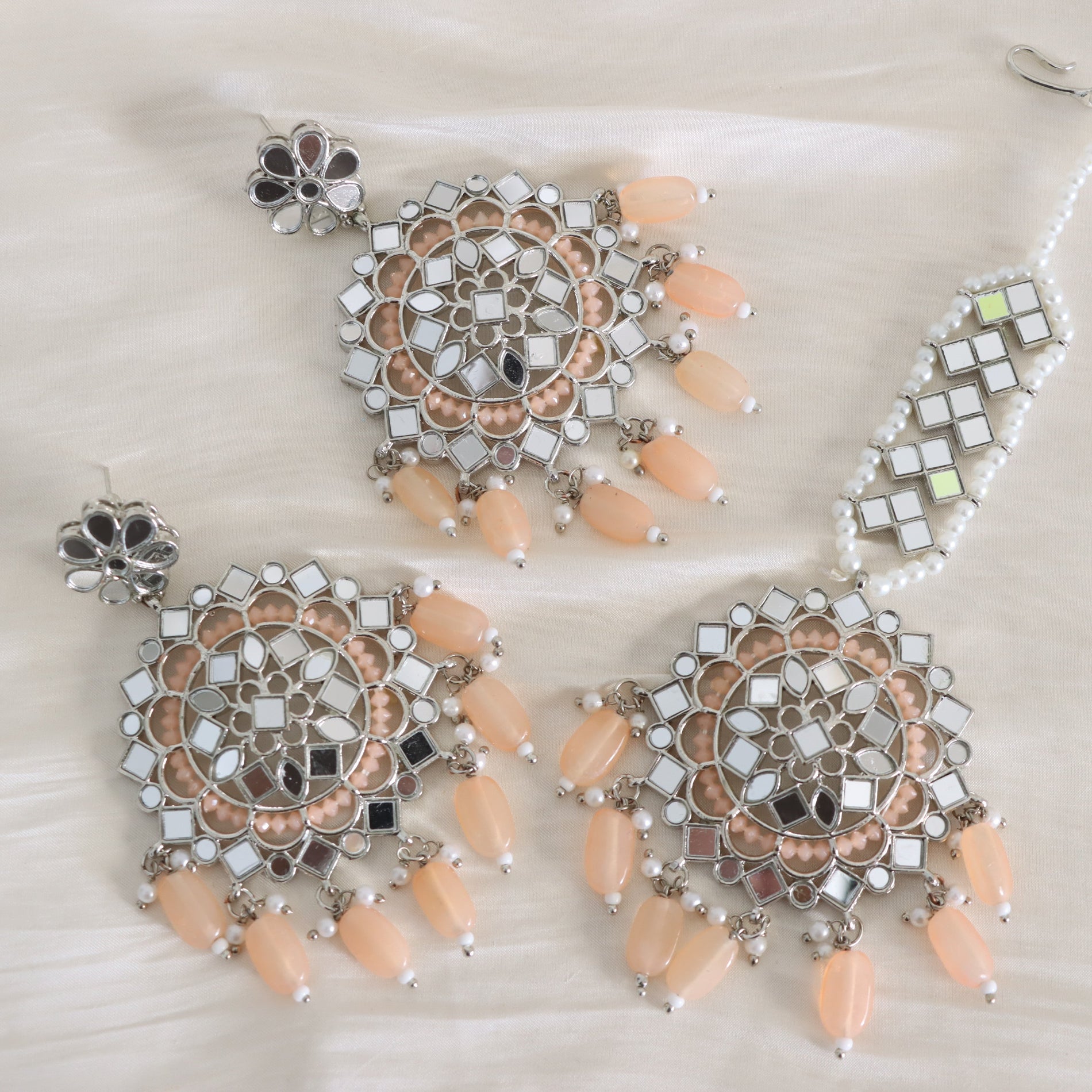 A traditional Indian bridal jewelry set featuring a pair of large chandelier earrings and a matching necklace, displayed on a soft white fabric background. The set includes silver-toned metalwork, white stones, peach-colored beads, and a strand of white faux pearls.