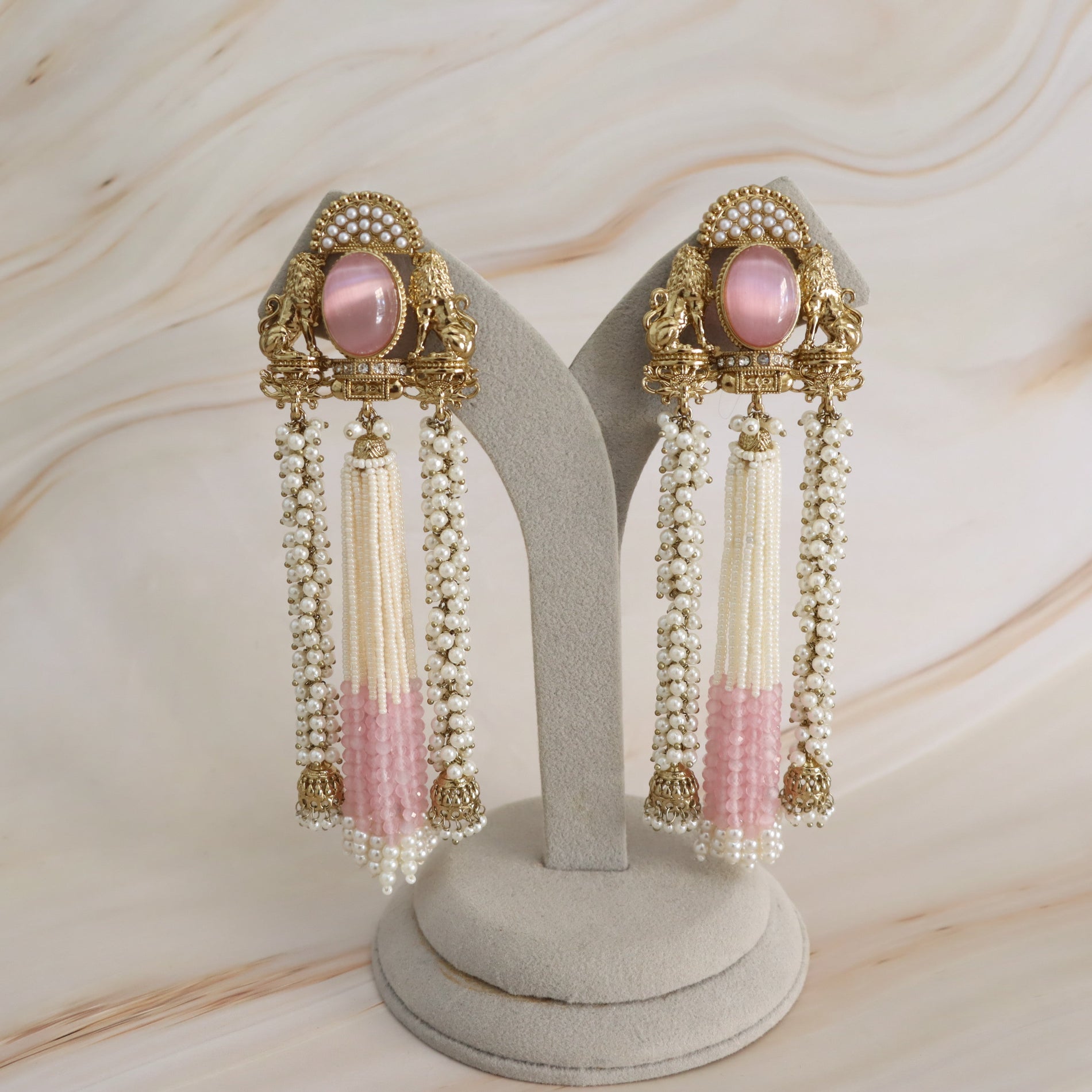 A pair of ornate gold-toned statement earrings with pink oval center stones and long dangling tassel strands, displayed on a gray jewelry stand against a light marble-patterned background.