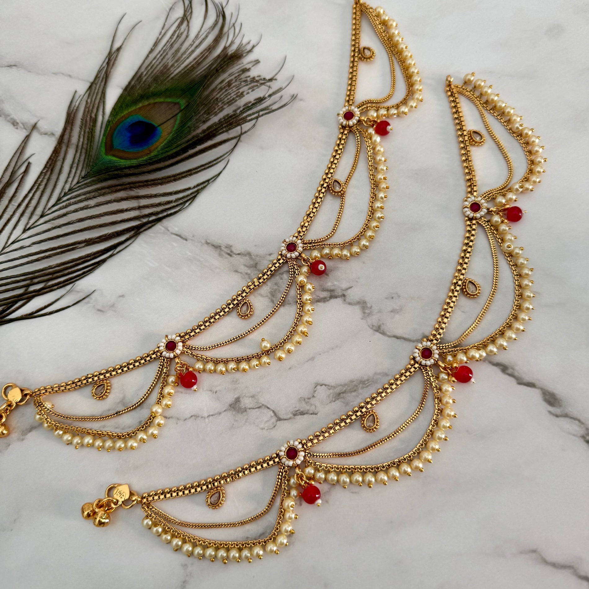 Gold anklet with pearls and red stones on a marble surface with a peacock feather.