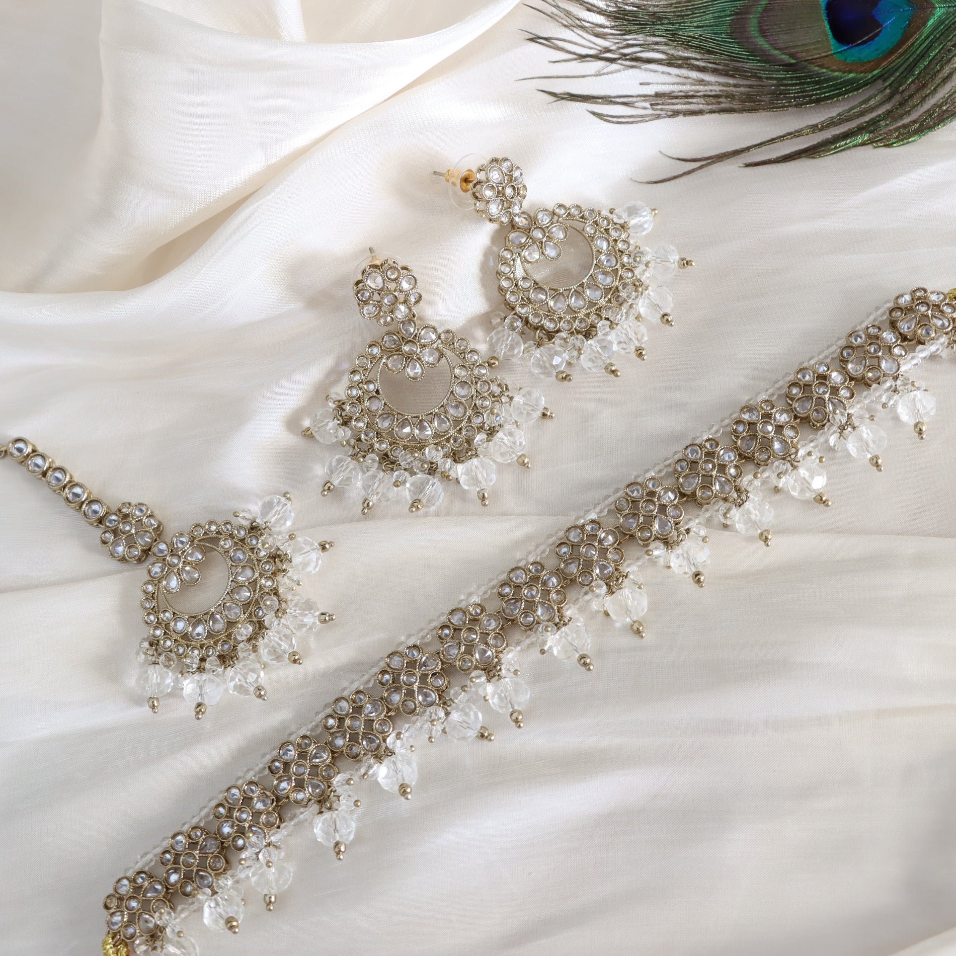 Beaded necklace and earrings on a white fabric background with a peacock feather.