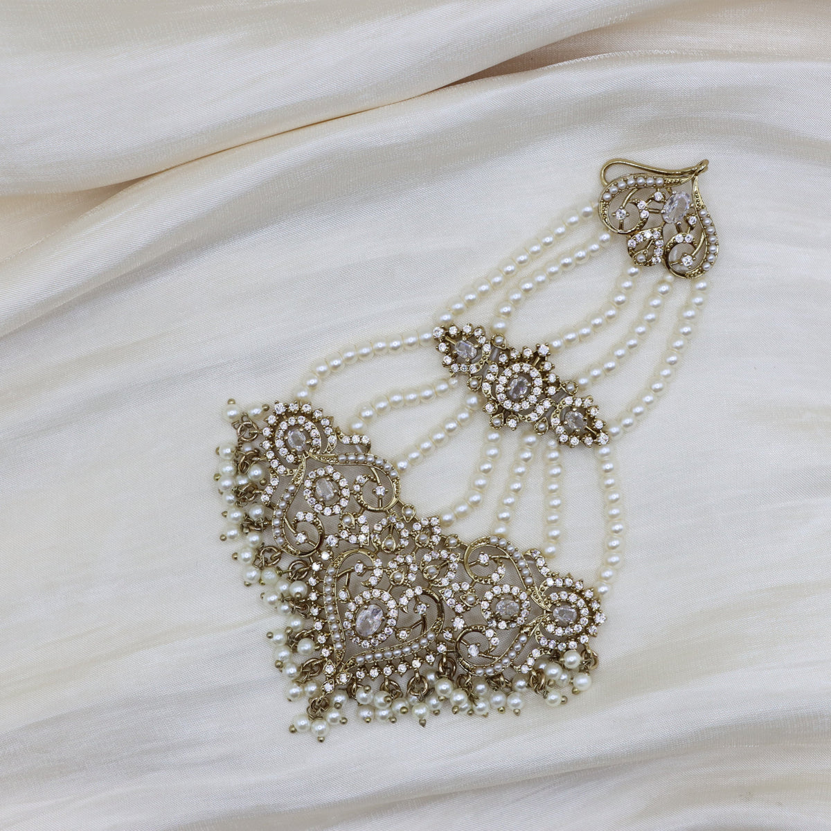 An ornate bridal headpiece with intricate gold-toned filigree, white faux pearls, and clear rhinestones, displayed on draped cream-colored fabric.