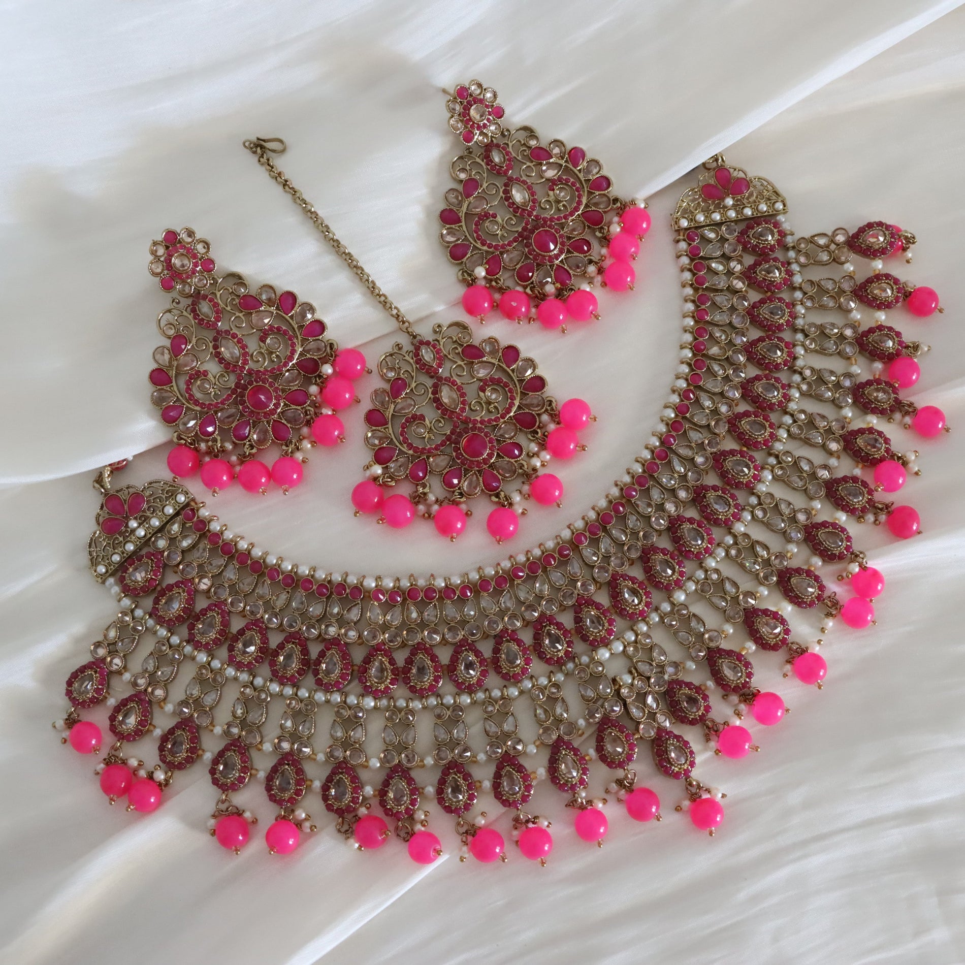 A traditional Indian jewelry set featuring a statement necklace, matching earrings, and a maang tikka, decorated with gold-plated filigree, pink gemstones, and bright pink bead accents, displayed on a white satin fabric.