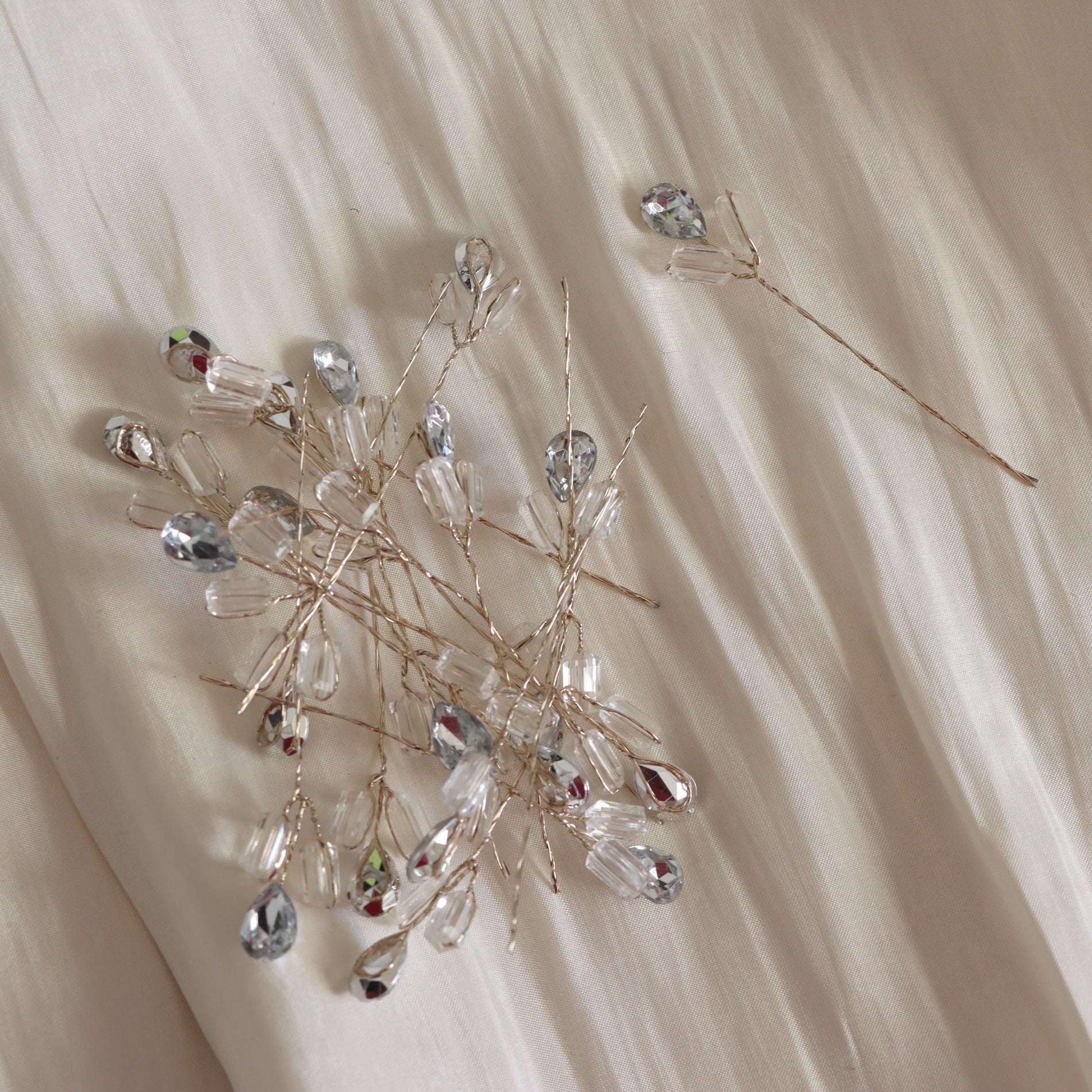 A set of delicate bridal hair accessories featuring clear teardrop and oval crystal beads on fine gold-toned wire branches, arranged on a draped cream fabric background.