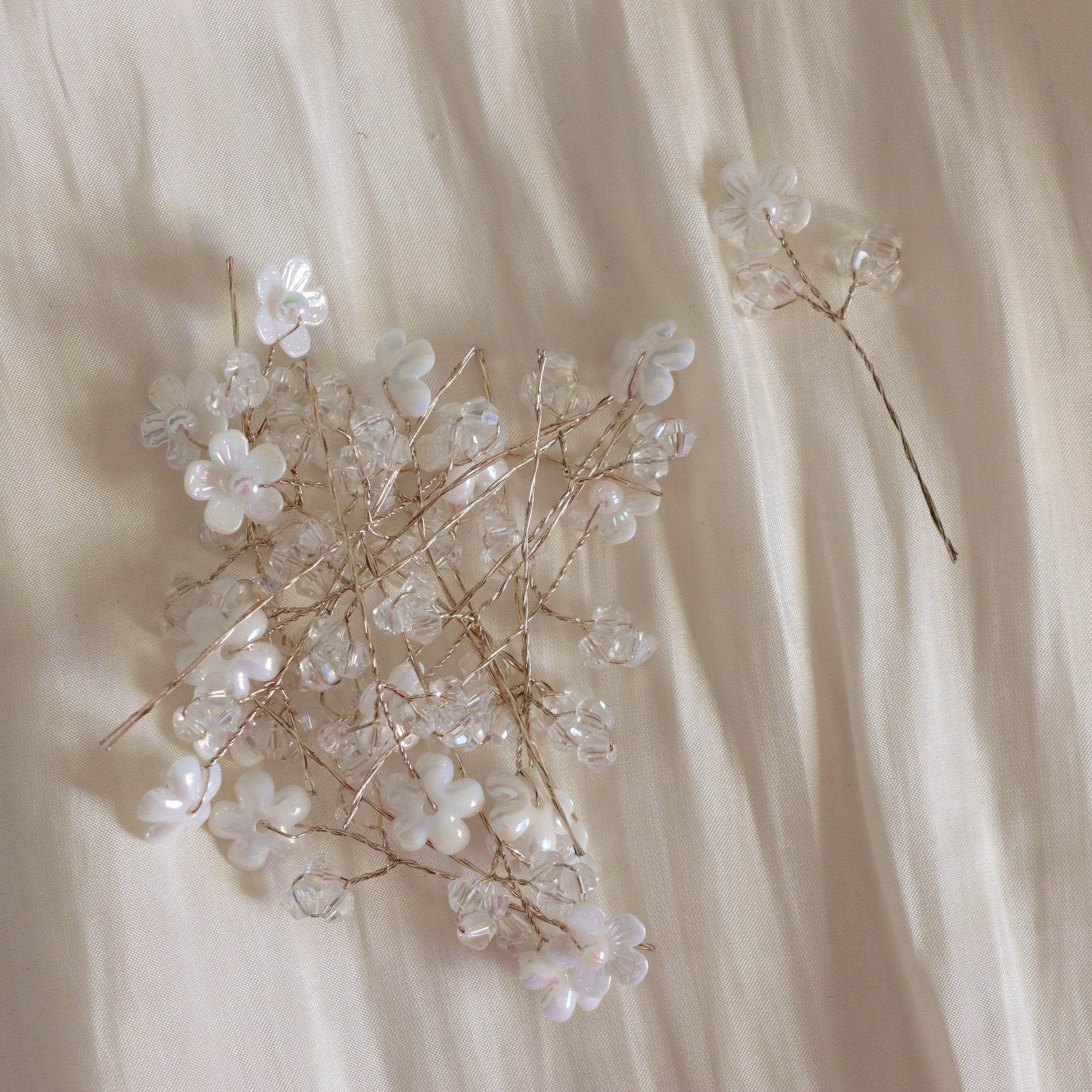 A delicate floral hair accessory made of clear crystal-like beads and small white flower elements on thin gold-toned wires, arranged on a soft cream fabric background.