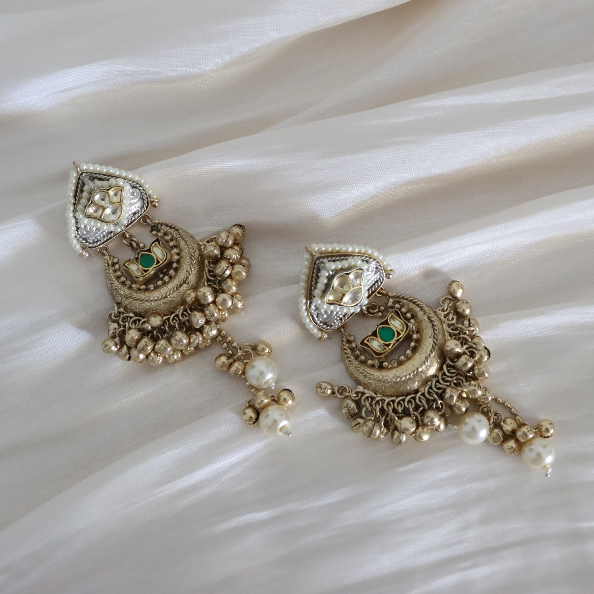 A pair of ornate gold-toned traditional Indian earrings with white pearl-like beads, small green stone accents, and silver-toned detailing, displayed on a draped white satin fabric background.
