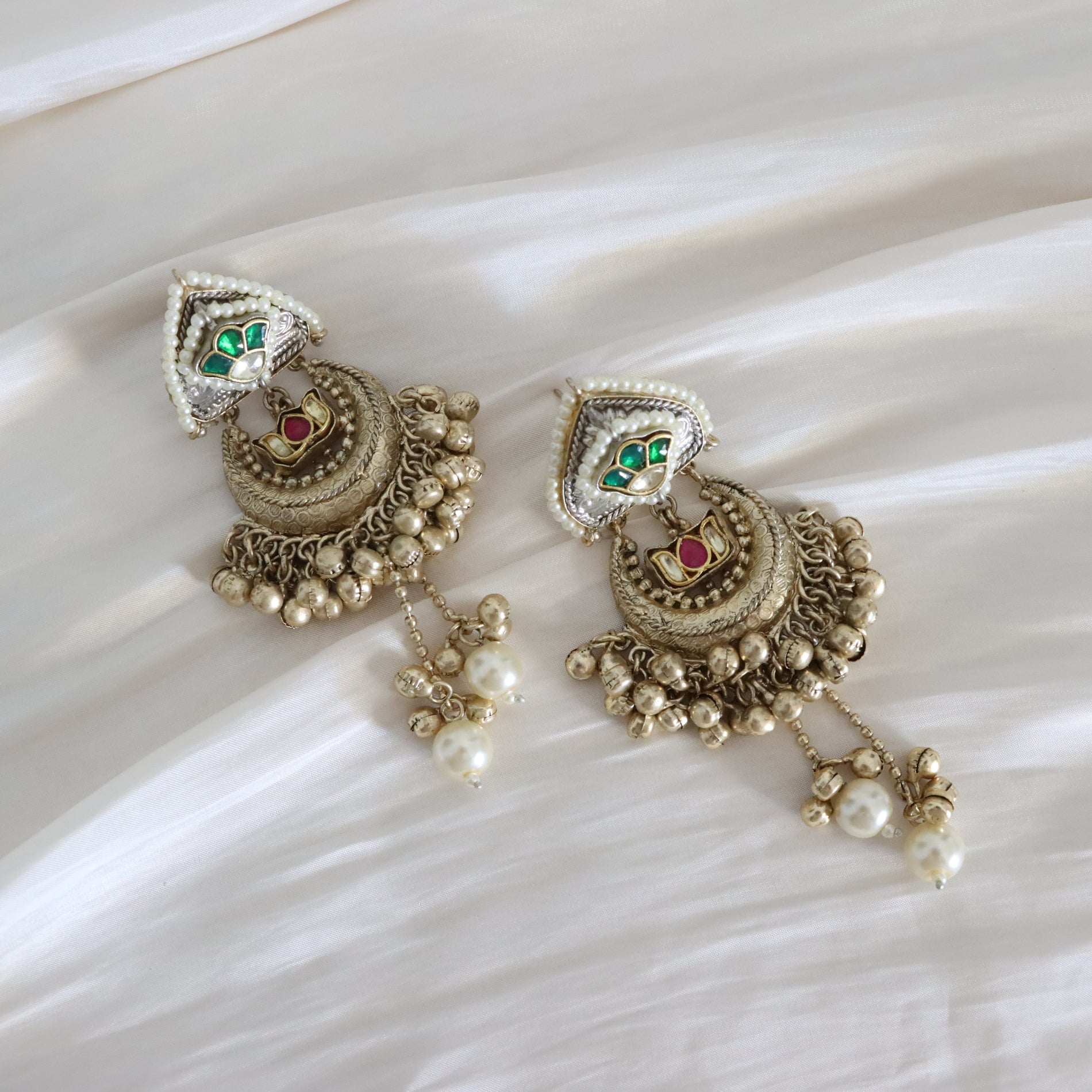 A pair of ornate Indian-style gold-toned chandelier earrings with white faux pearls, green and red enamel details, and silver-toned accents, displayed on a draped white fabric background.