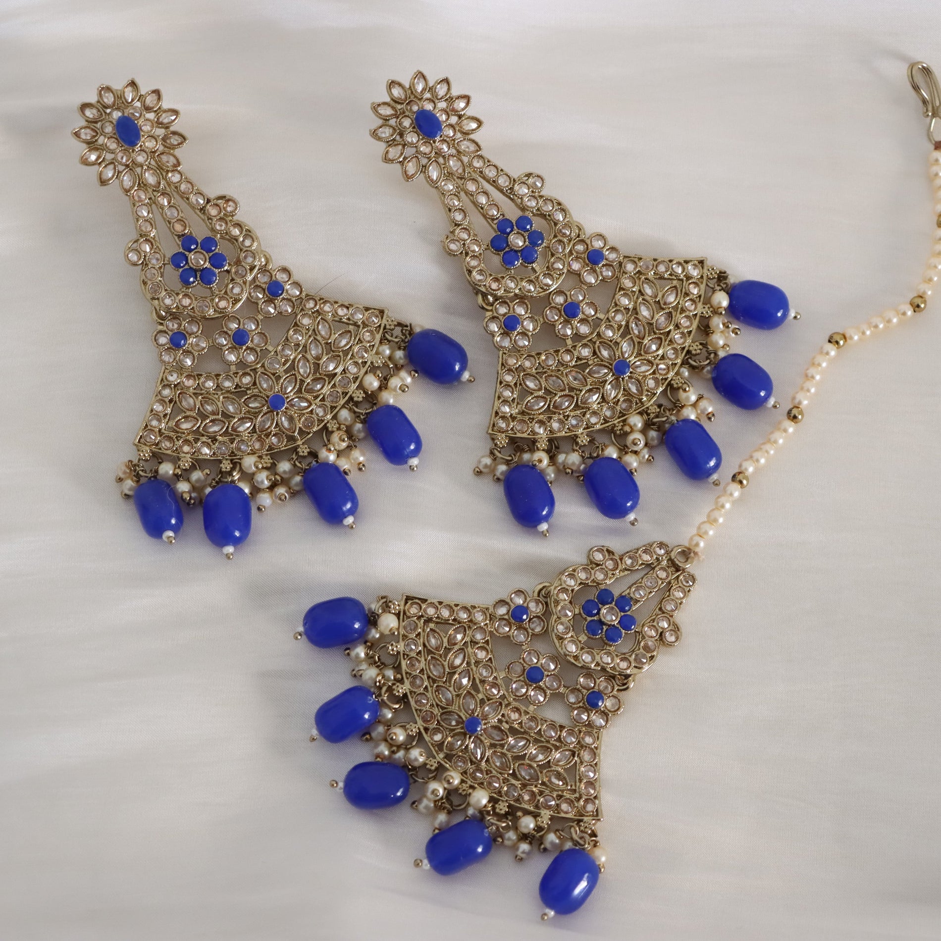 Gold-toned traditional Indian jewelry set featuring a maang tikka and matching necklace with blue stone accents and pearl-like bead details, displayed on a white fabric background.
