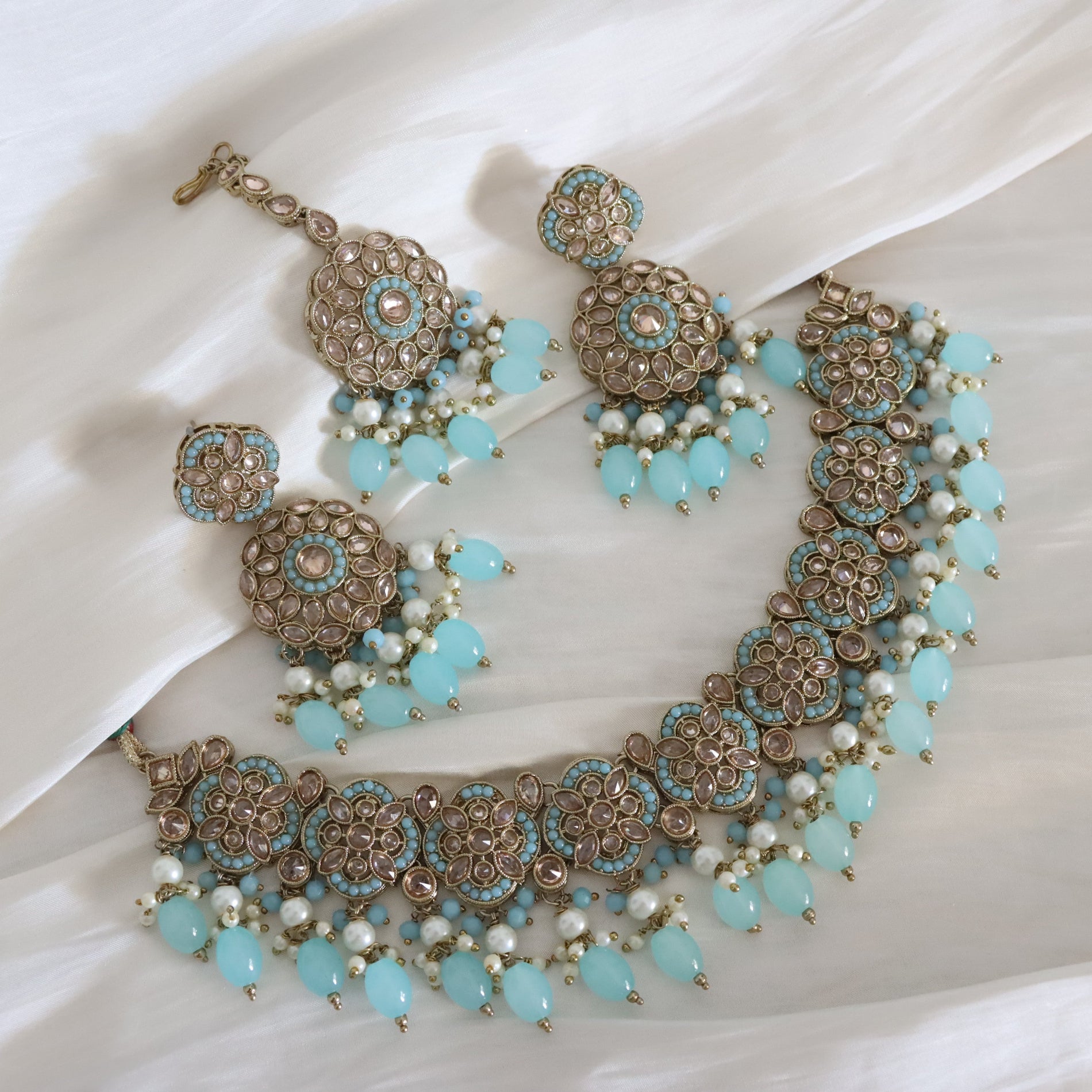 An ornate Indian-style jewelry set featuring a statement necklace and matching earrings, decorated with light blue beads, white faux pearls, and intricate gold-toned filigree, displayed on a draped white fabric background.