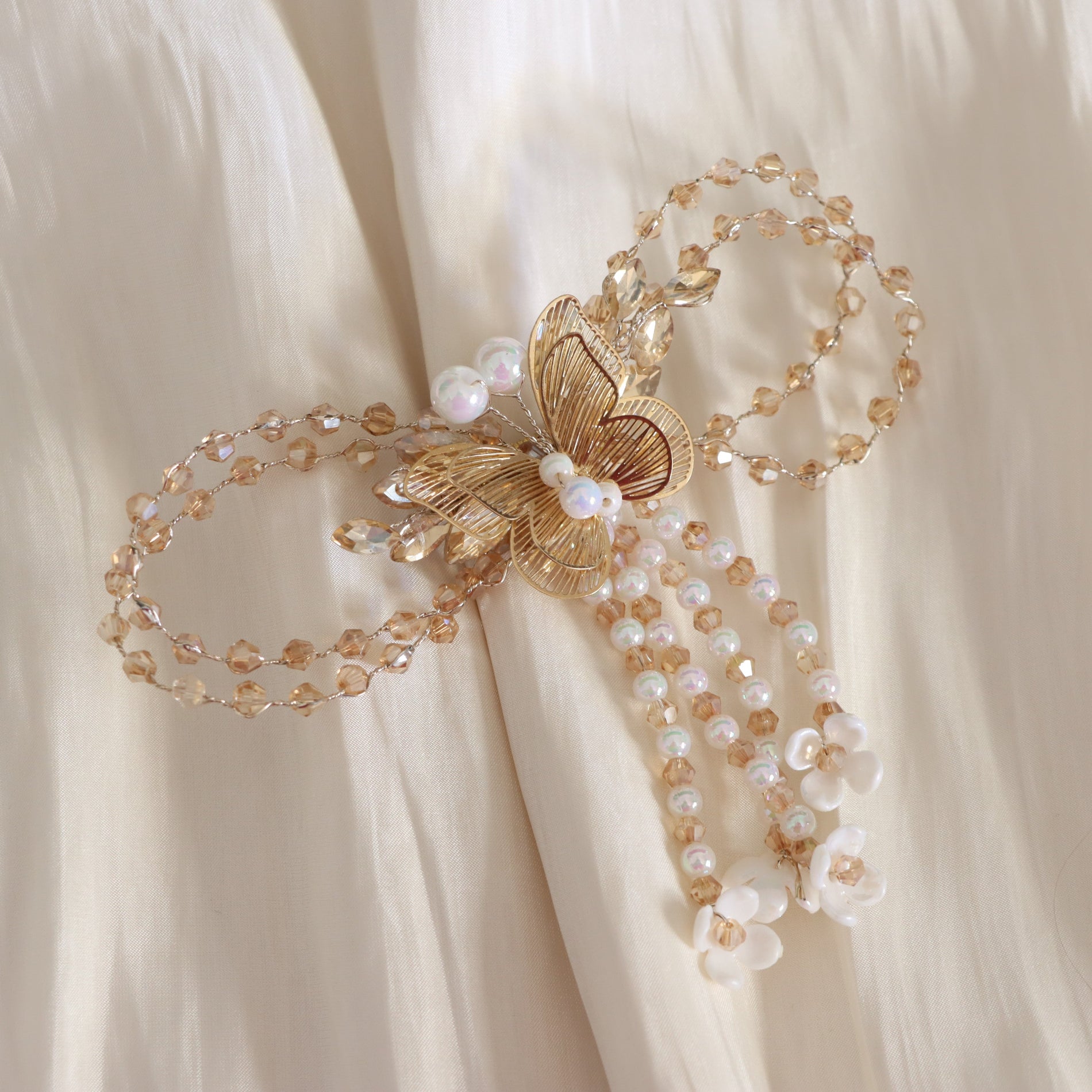 A delicate gold butterfly hair accessory with pearl-like beads, clear faceted beads, and small white flower charms, displayed on draped cream fabric.