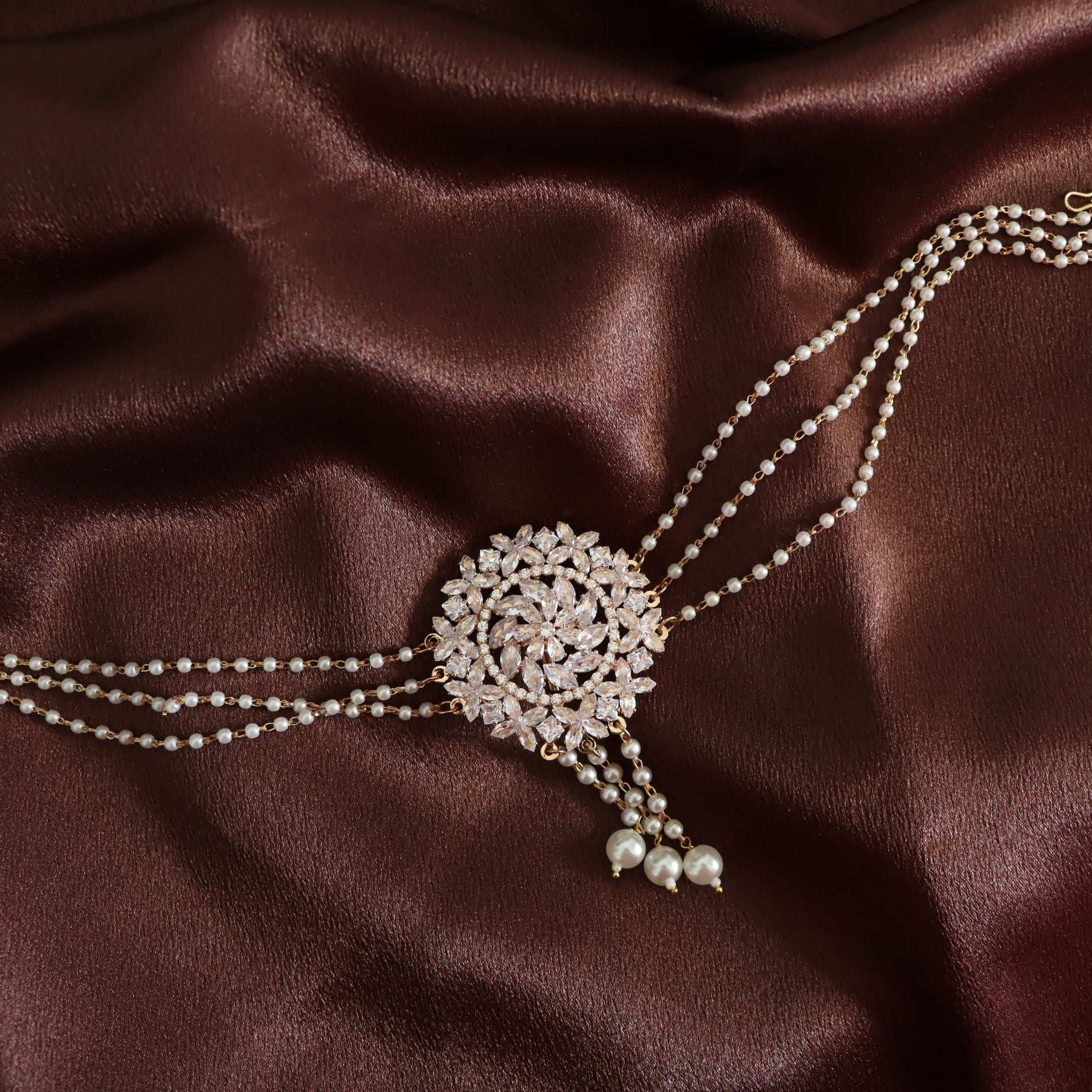 A double-strand pearl choker necklace with a large, ornate round pavé centerpiece and dangling pearl beads, displayed on a draped dark brown satin fabric.