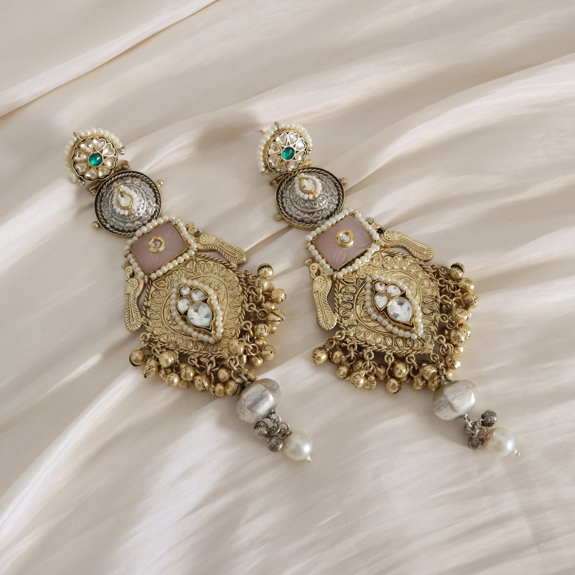 A pair of ornate Indian-style gold-toned dangle earrings with pink enamel, white pearls, and small green stones, displayed on a draped cream satin fabric background.
