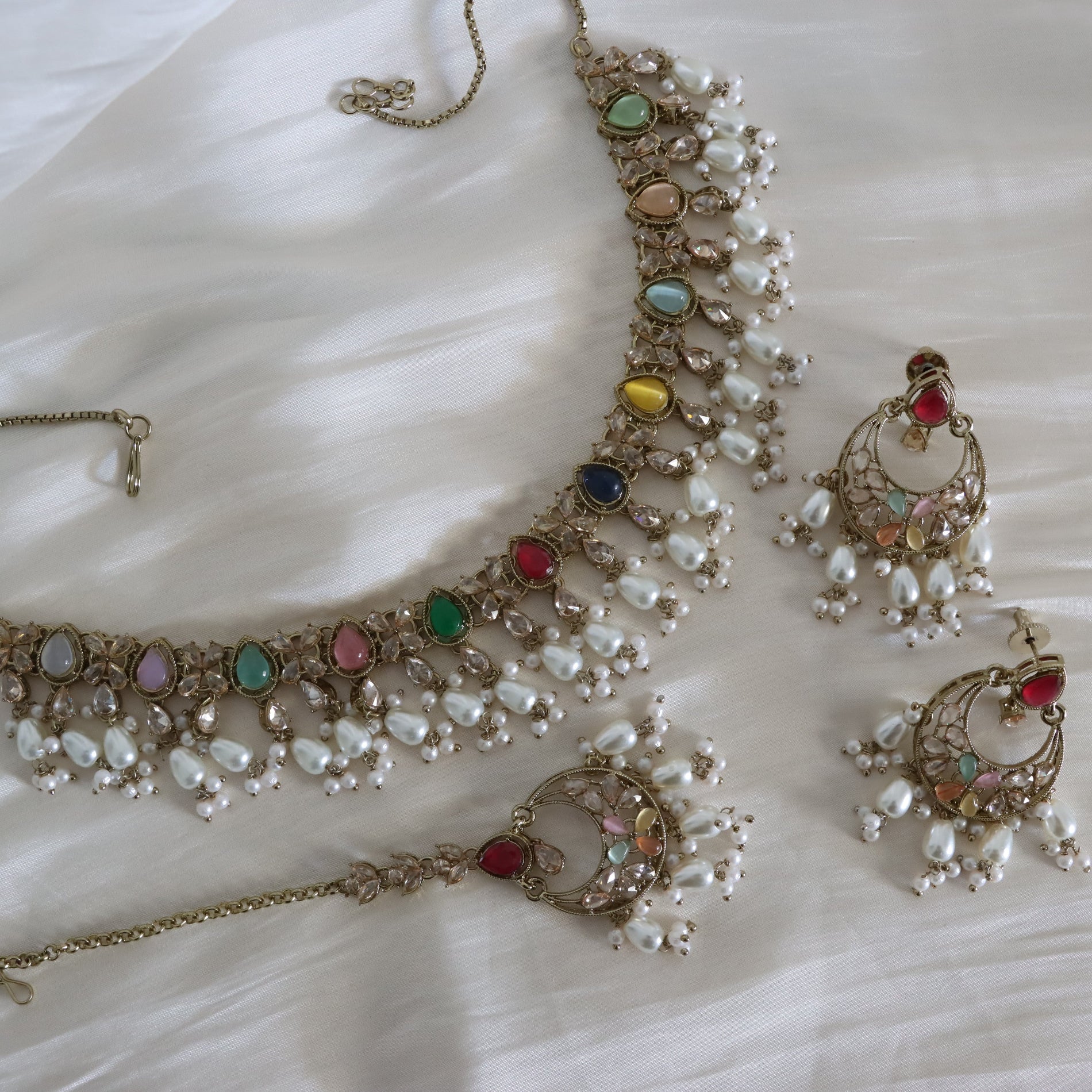 An ornate Indian ethnic jewelry set featuring a necklace, earrings, and a bracelet, decorated with white pearls and multicolored gemstones in gold-plated settings, laid out on a white fabric background.