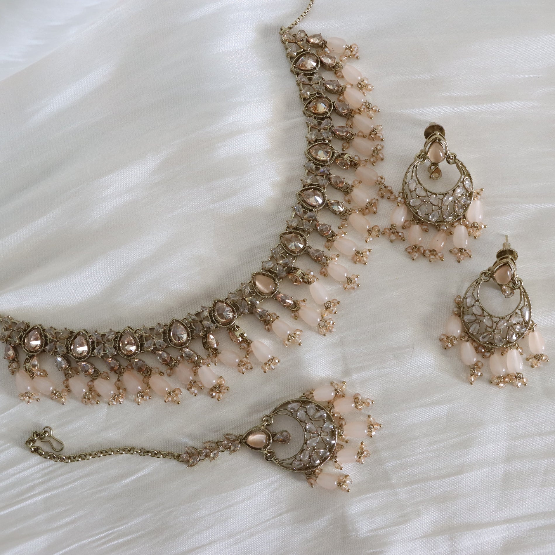 A traditional Indian jewelry set featuring a necklace, earrings, and a bracelet in rose gold with light pink beads, displayed on a white textured fabric background.