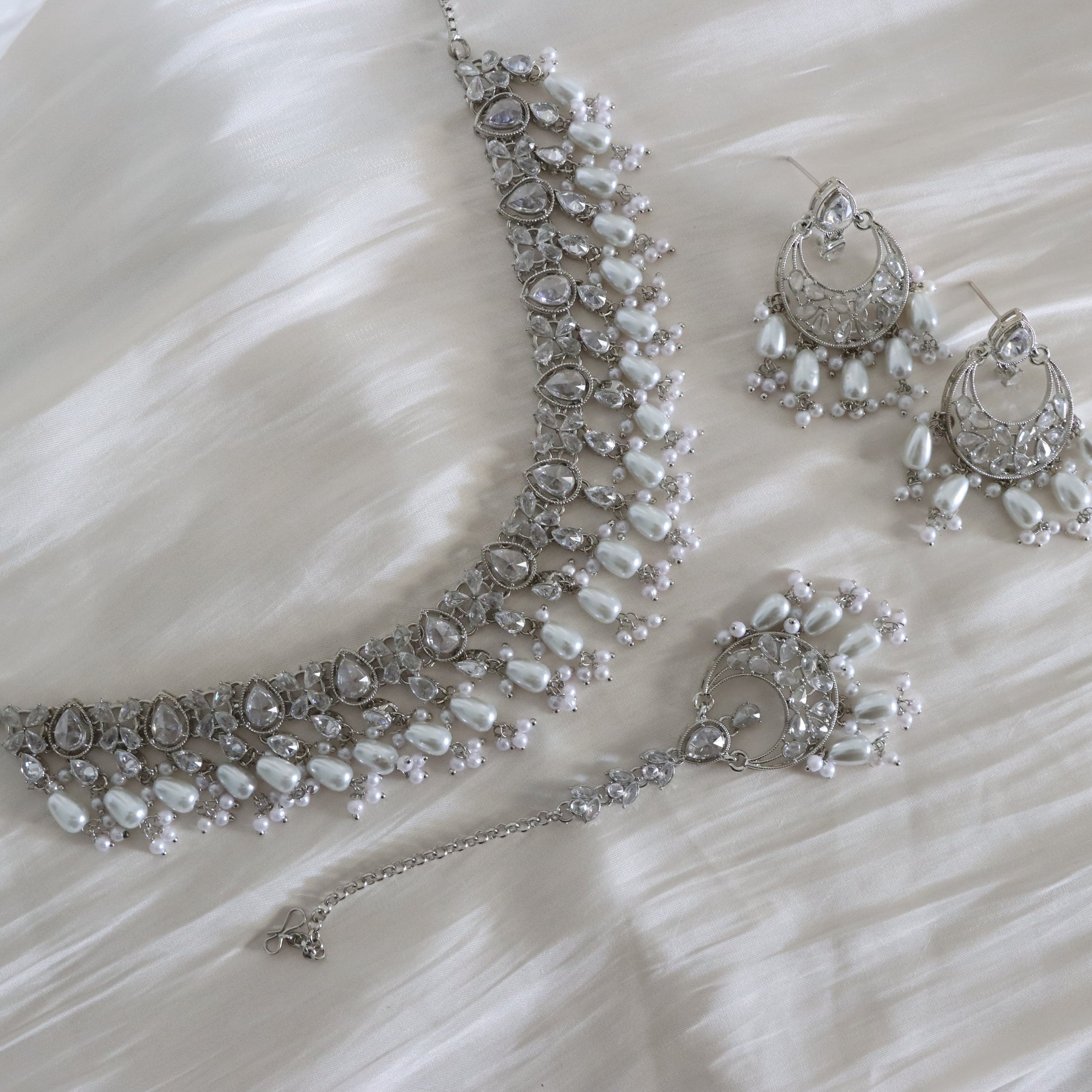 Silver-toned bridal jewelry set featuring a pearl and crystal necklace, matching earrings, and a dangling bracelet, arranged on a white faux fur surface.