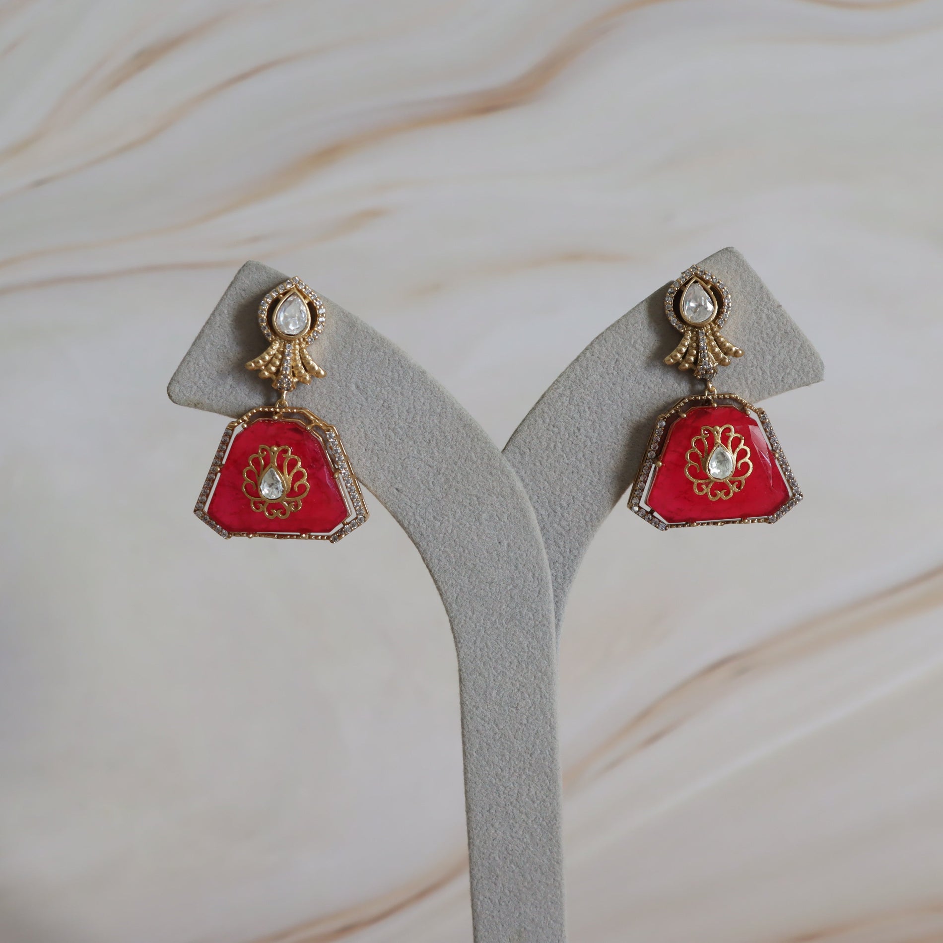 Pair of red and gold earrings on a gray stand with a beige background