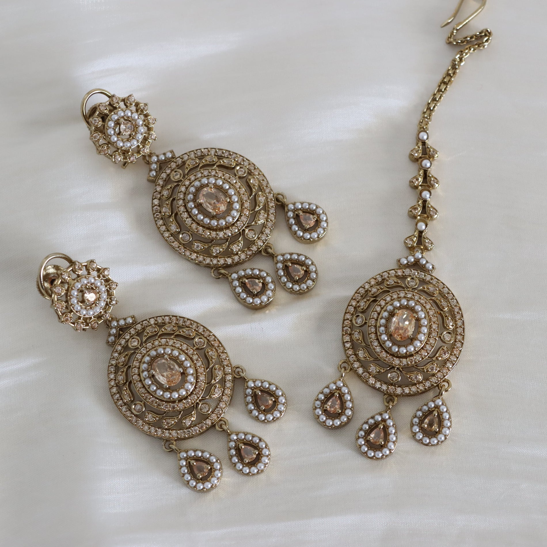 Gold-toned traditional Indian jewelry set including a choker necklace, matching earrings, and a maang tikka, embellished with gold-plated circular motifs, small white faux pearls, and central peach-colored stones, displayed on a white satin fabric.