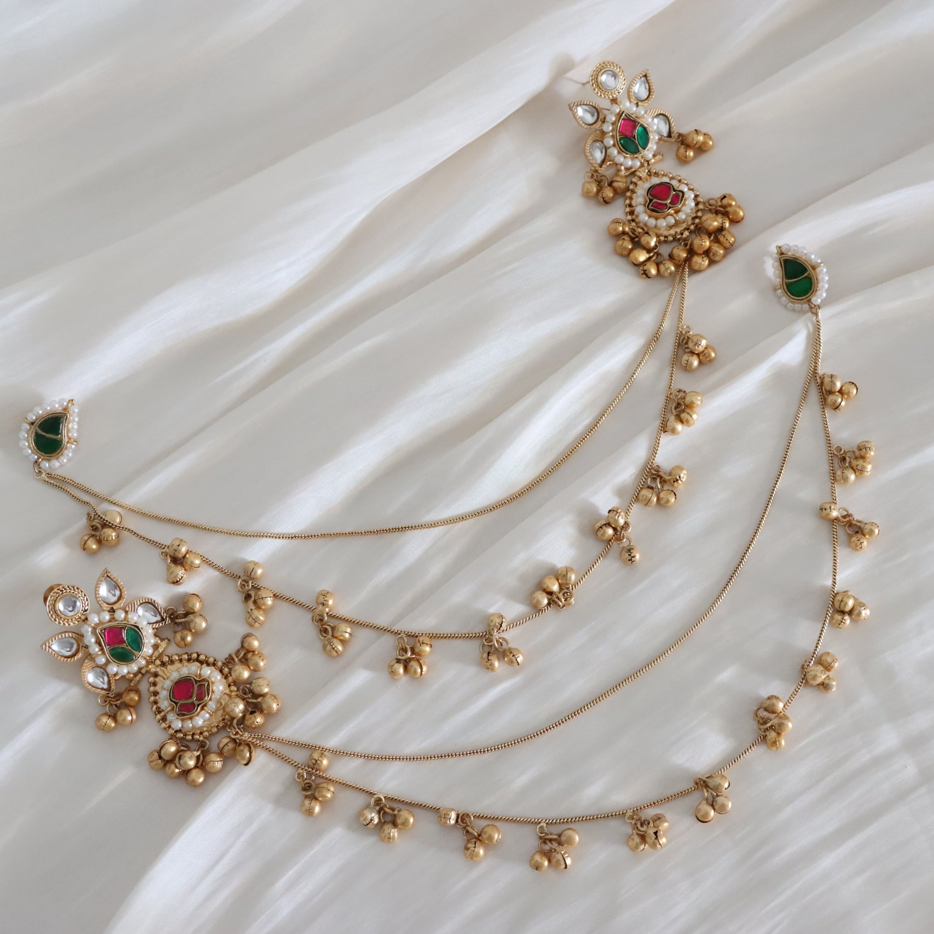 Gold-toned traditional Indian jewelry set including a long chain necklace with dangling floral motifs, a matching headpiece, and a small matching earring, decorated with green and red stones and white crystal-like accents, displayed on a white draped fabric background.