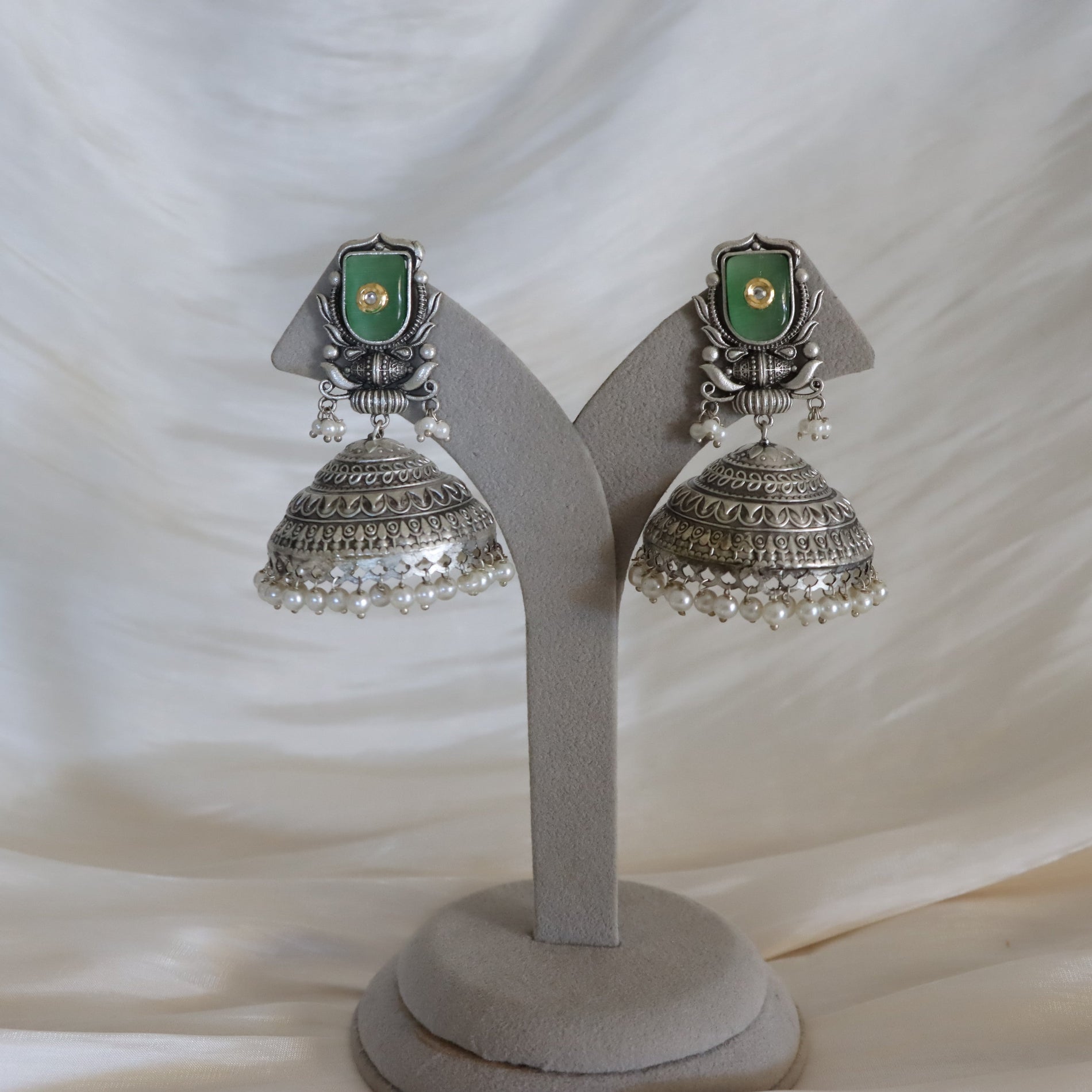 A pair of traditional Indian silver jhumka earrings with green stone inlays and dangling pearl bead borders, displayed on a gray velvet jewelry stand against a draped white fabric background.