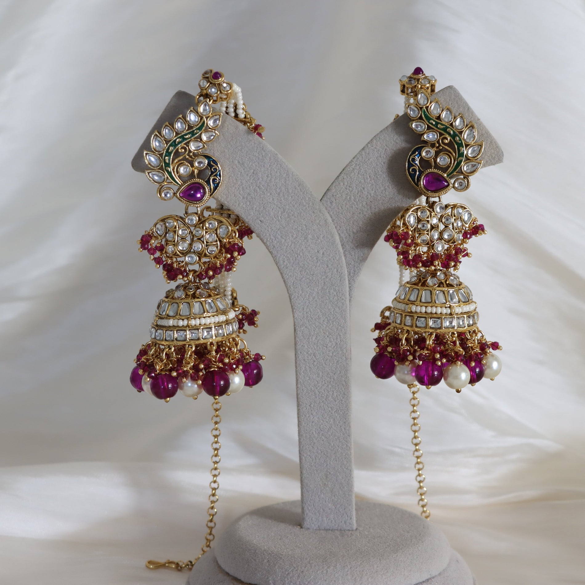 A pair of ornate gold-toned Indian jhumka earrings with white stones, purple gemstone accents, and dangling pearl-like beads, displayed on a gray jewelry stand against a white fabric background.