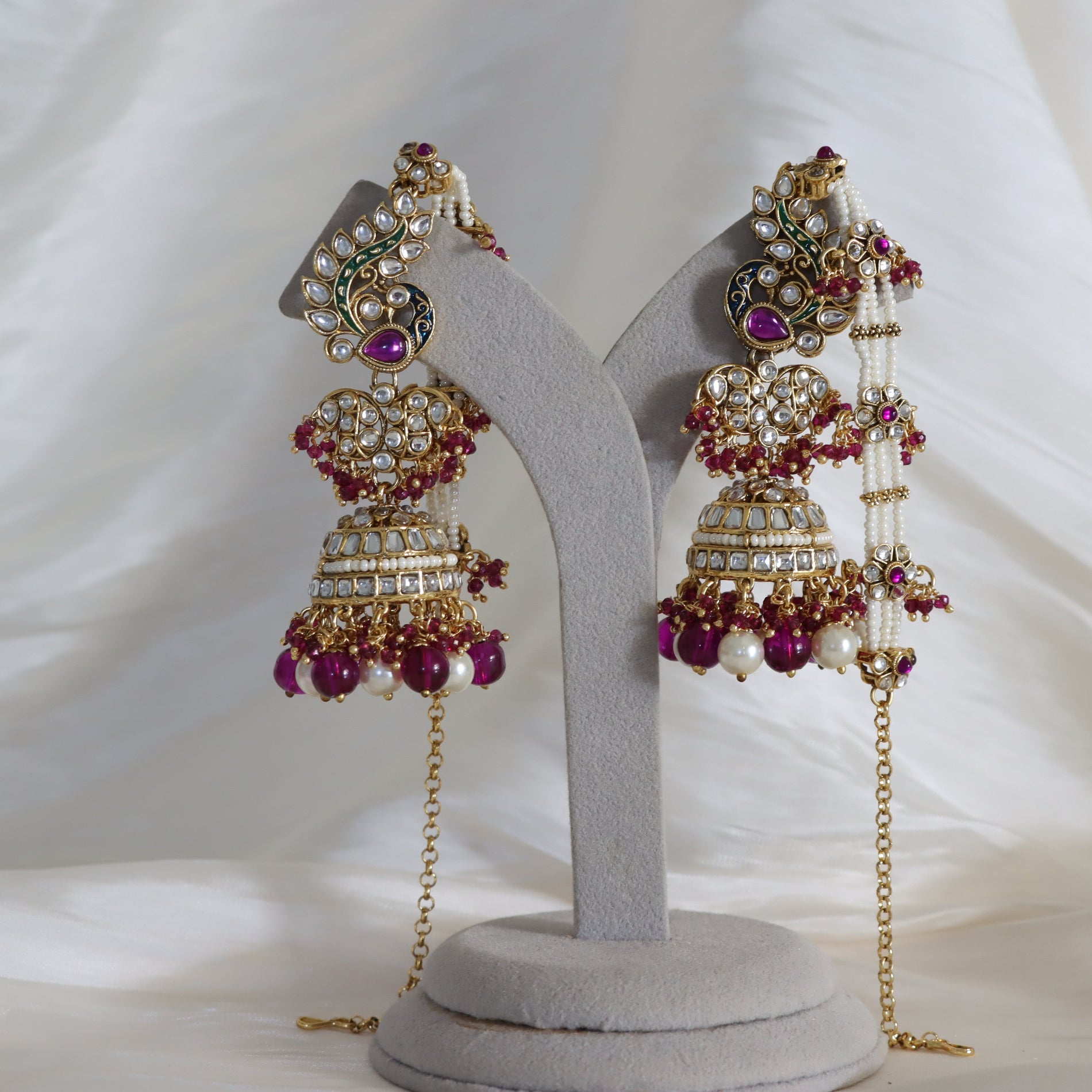 A pair of ornate gold-toned Indian jhumka earrings with white pearls, clear crystals, and deep purple gemstones, displayed on a gray velvet jewelry stand against a white draped fabric background.