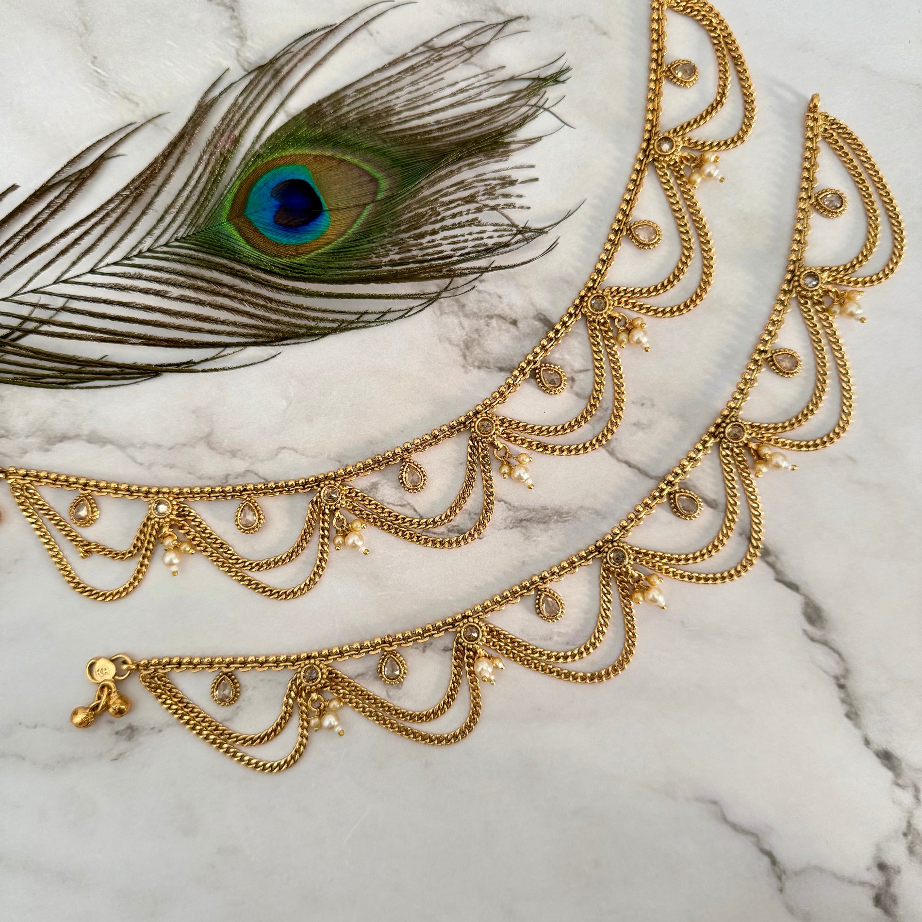 Gold anklet on a marble surface with a peacock feather