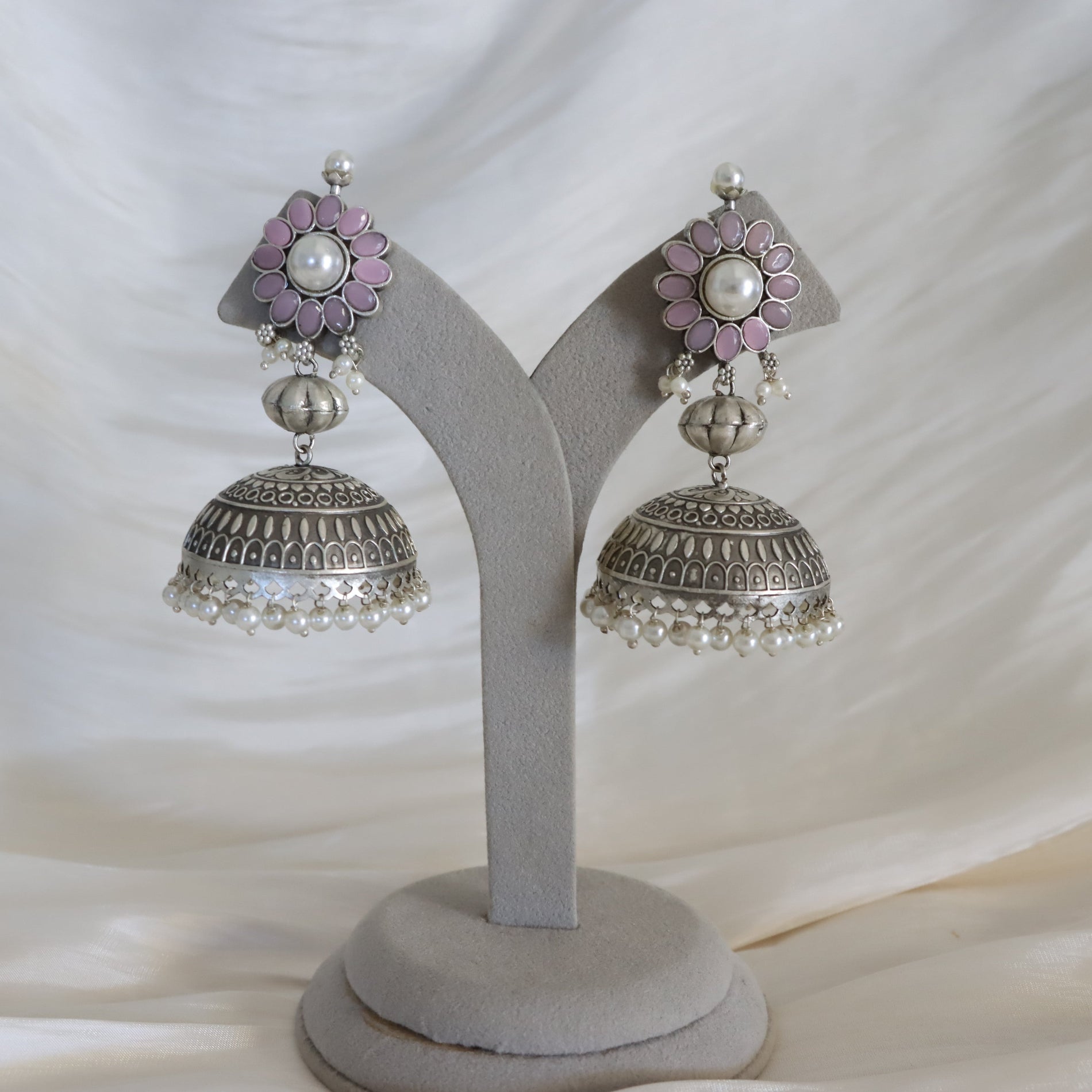 A pair of silver jhumka earrings with pink floral accents and white pearl details, displayed on a gray velvet jewelry stand against a draped white fabric background.