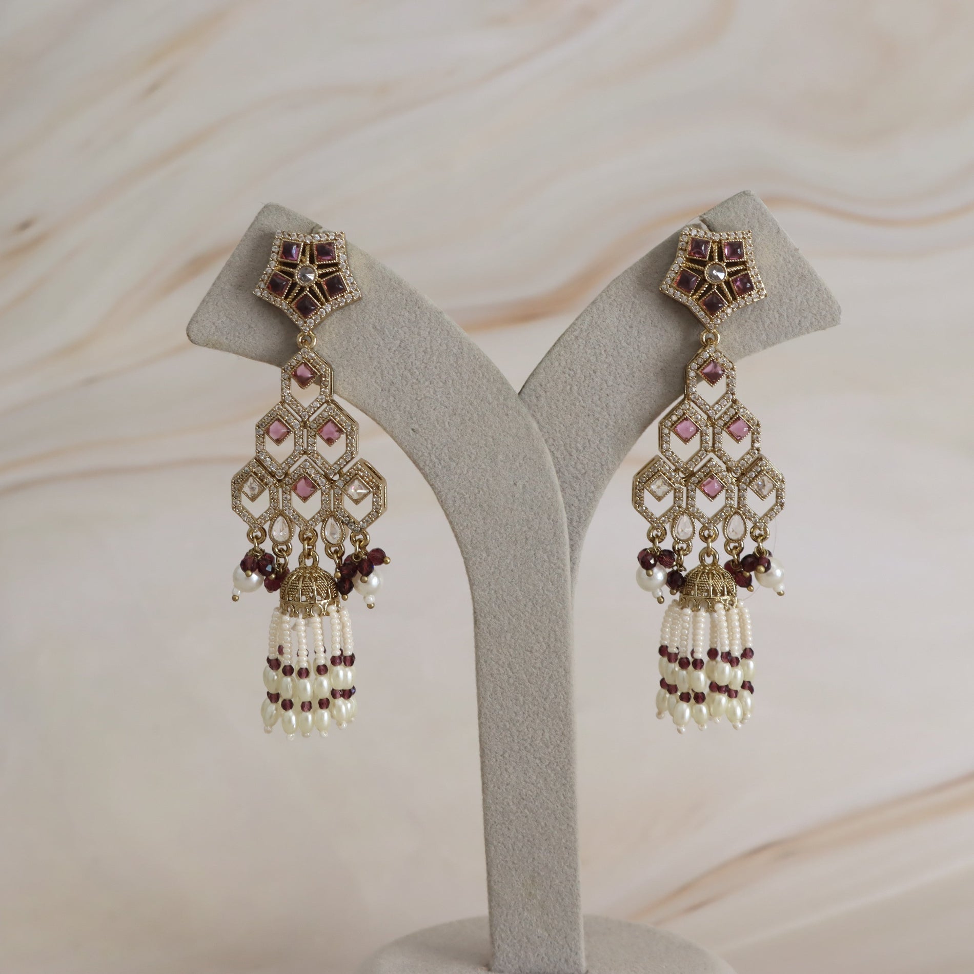 A pair of ornate gold-toned Indian-style dangle earrings with pink and dark red stones and white pearl tassels, displayed on a gray velvet jewelry stand against a light beige marble background.