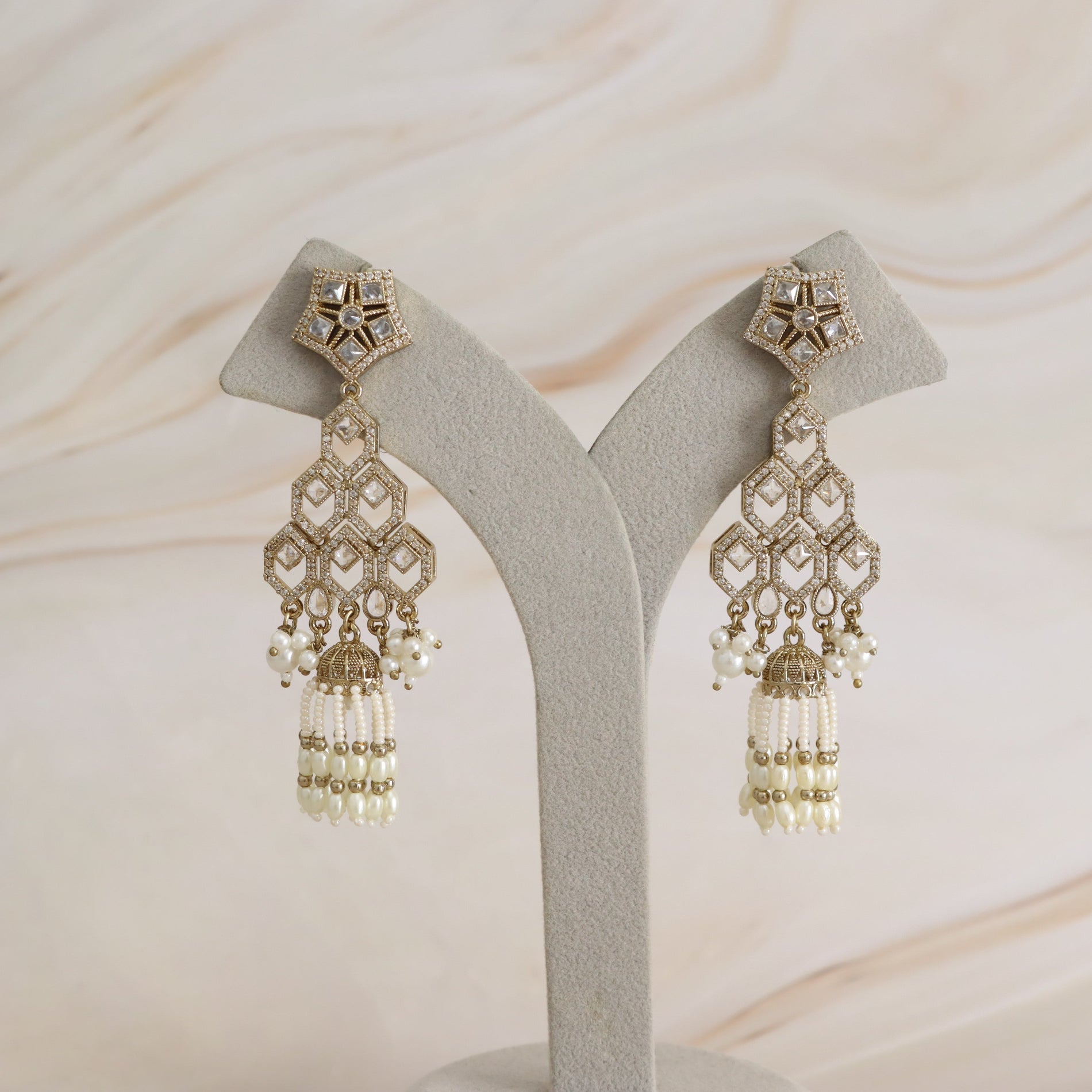 A pair of ornate gold-toned statement earrings displayed on a light gray velvet jewelry stand against a soft beige marble background. The earrings feature intricate geometric filigree, small clear stones, and dangling pearl-like bead tassels.