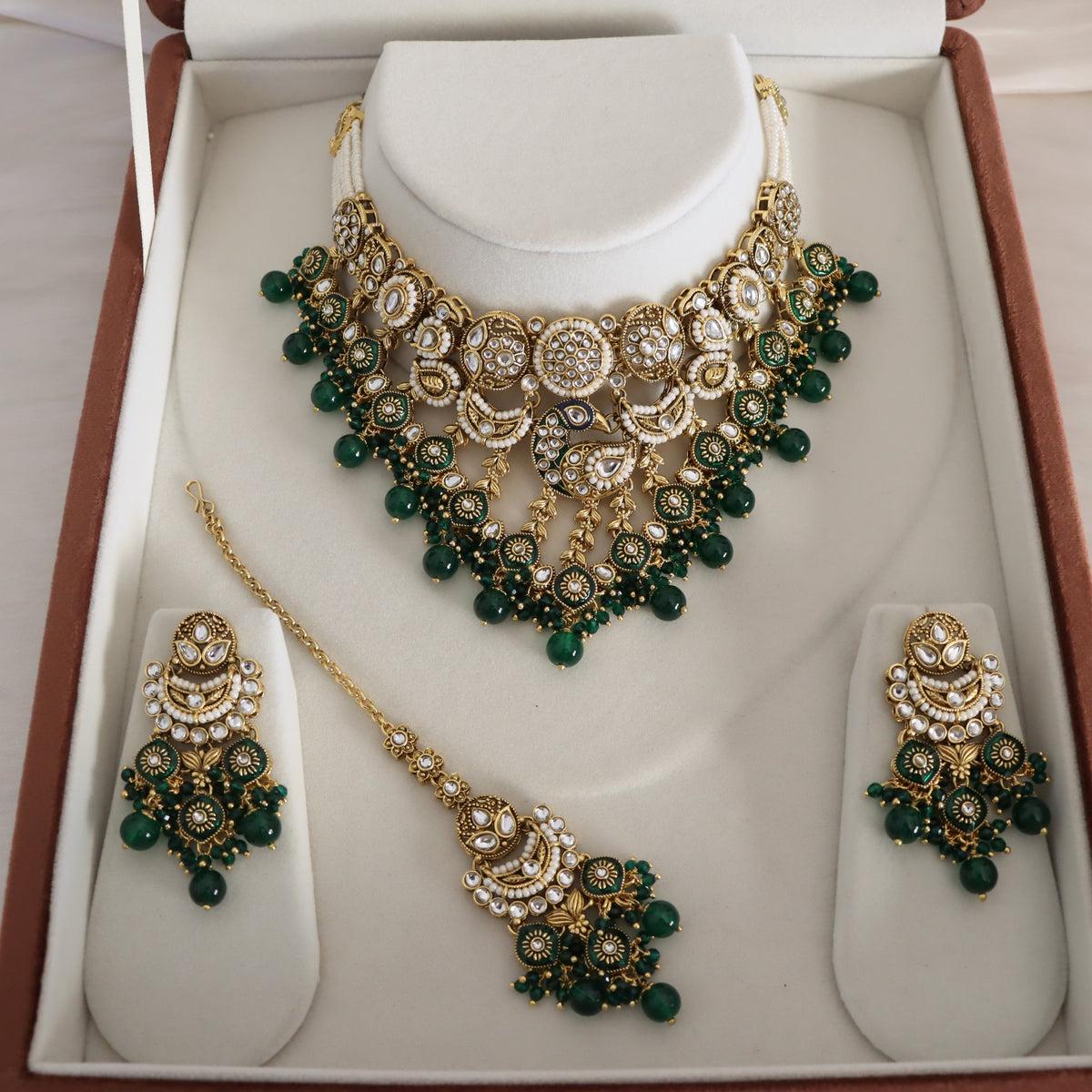 Gold and green traditional Indian jewelry set displayed in a white jewelry box, including a statement necklace, matching earrings, and a maang tikka, with clear stones and green beads.
