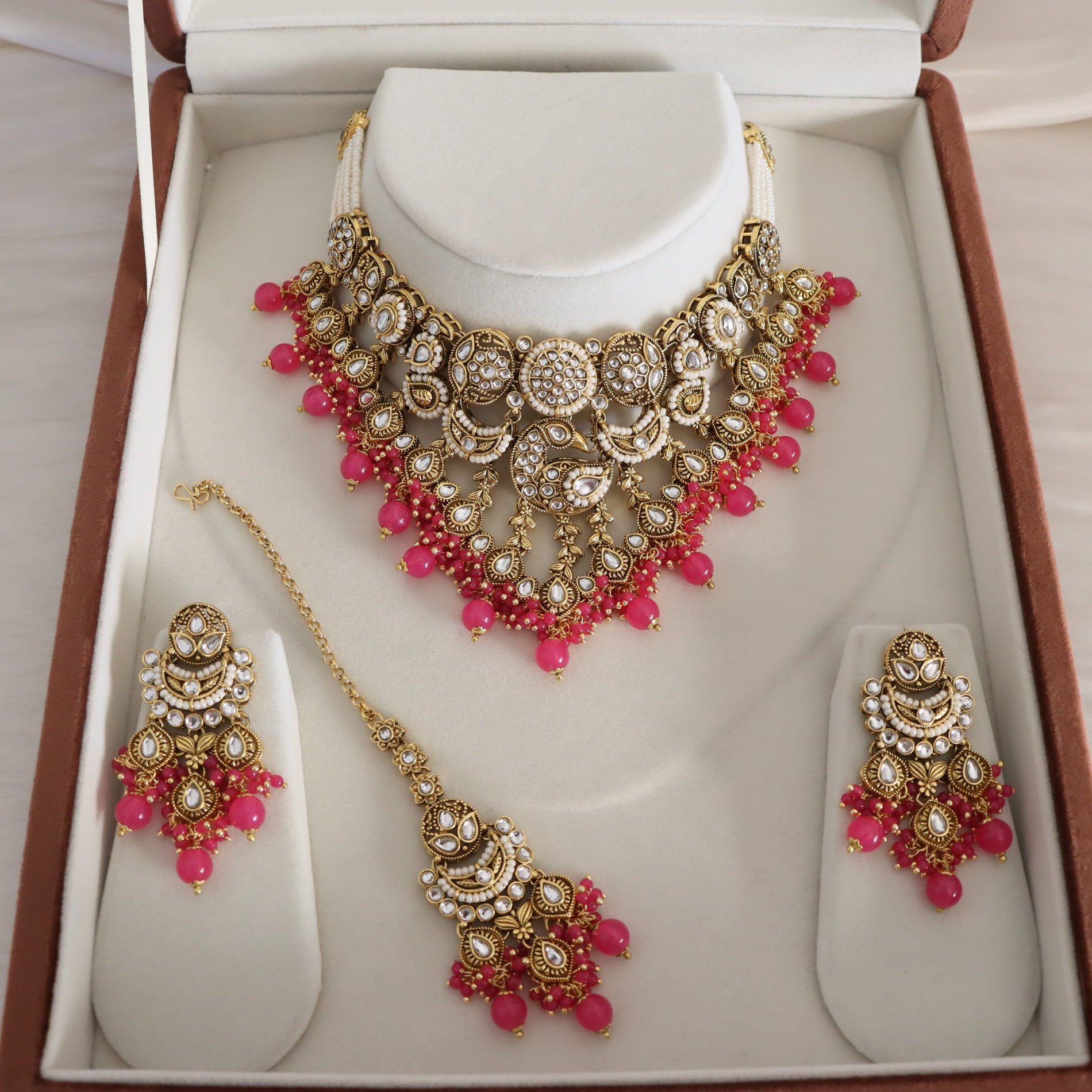 An ornate Indian traditional jewelry set displayed inside an open presentation box. The set includes a gold-plated necklace with white stones and pink beads, matching drop earrings, and a maang tikka, all arranged on white display stands.