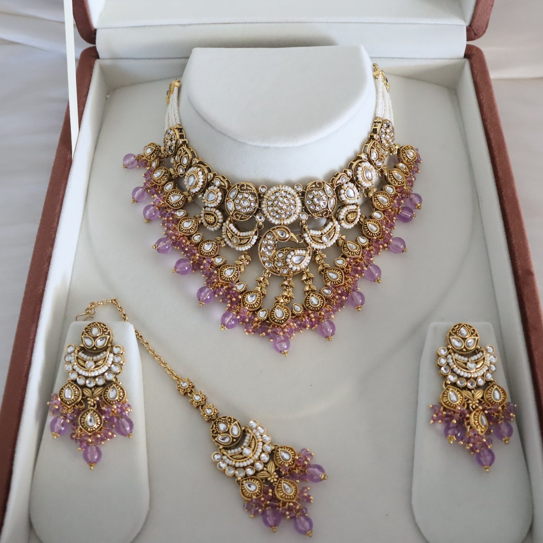 An ornate gold-toned traditional jewelry set displayed inside an open presentation box. The set includes a statement necklace on a white neck form, matching earrings on standees, and a dangling maang tikka, decorated with white faux pearls, gold filigree, and purple bead accents.