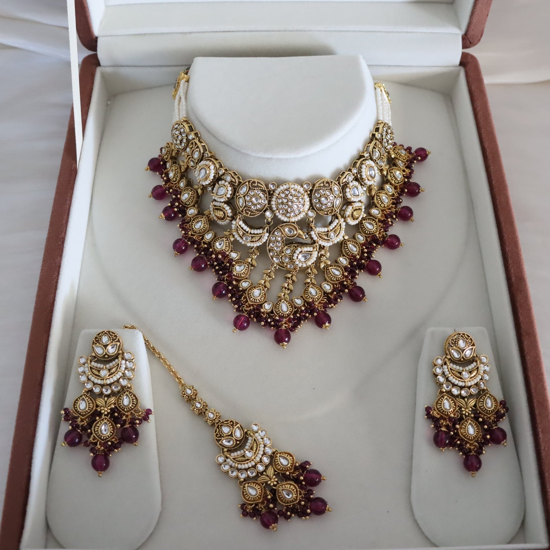 Gold-toned traditional Indian jewelry set displayed in an open presentation box, including a choker necklace, matching earrings, and a chain with a pendant. The pieces feature gold-plated metal, white faux pearls, and deep red gemstone accents.