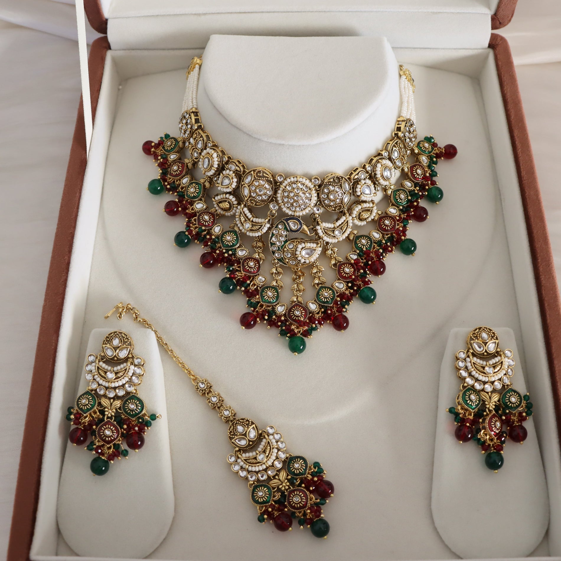 A traditional Indian gold jewelry set displayed inside an open presentation box. The set includes a statement necklace, matching earrings, and a pendant, all decorated with gold-plated metal, clear stones, and green and red beads.