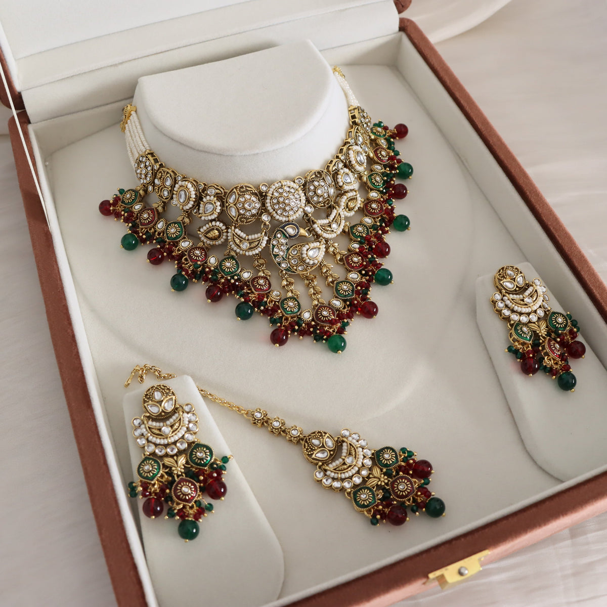 A traditional Indian jewelry set including a statement necklace, matching earrings, and a pendant, displayed on white velvet stands inside a brown presentation box.