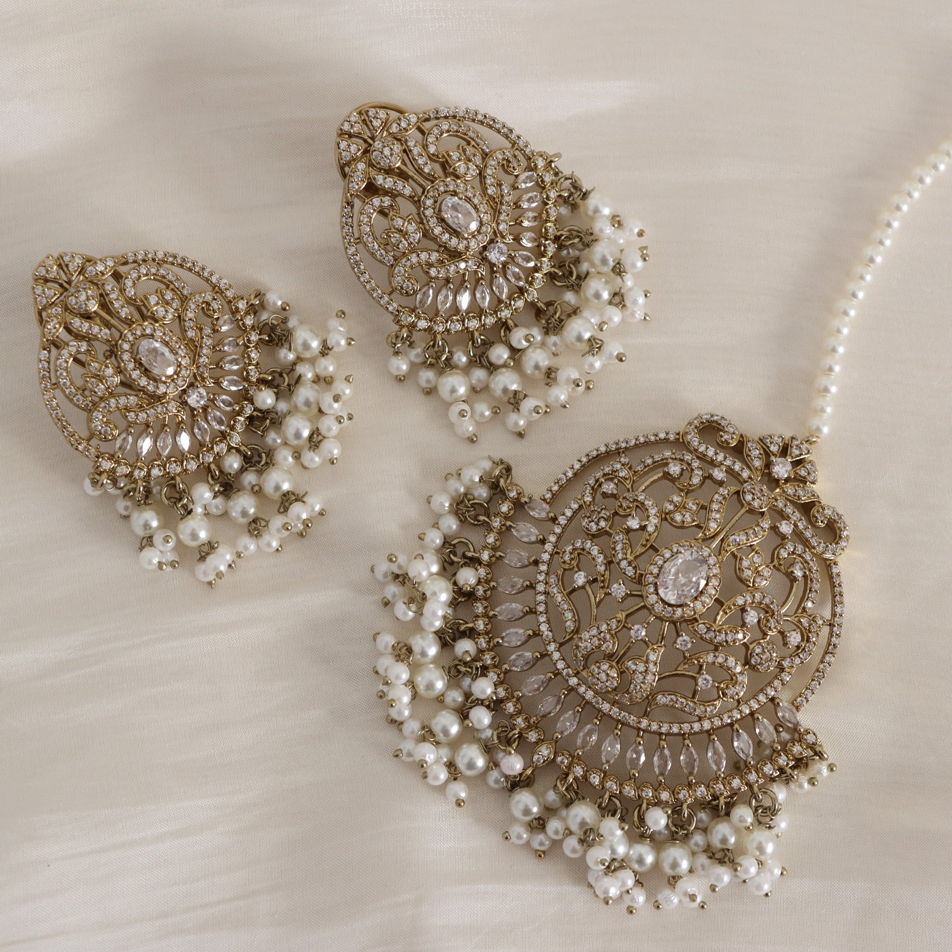 A traditional Indian bridal jewelry set featuring a gold-plated necklace with intricate filigree work, white faux pearls, and clear crystals, accompanied by matching earrings and a pearl necklace on a soft cream fabric background.