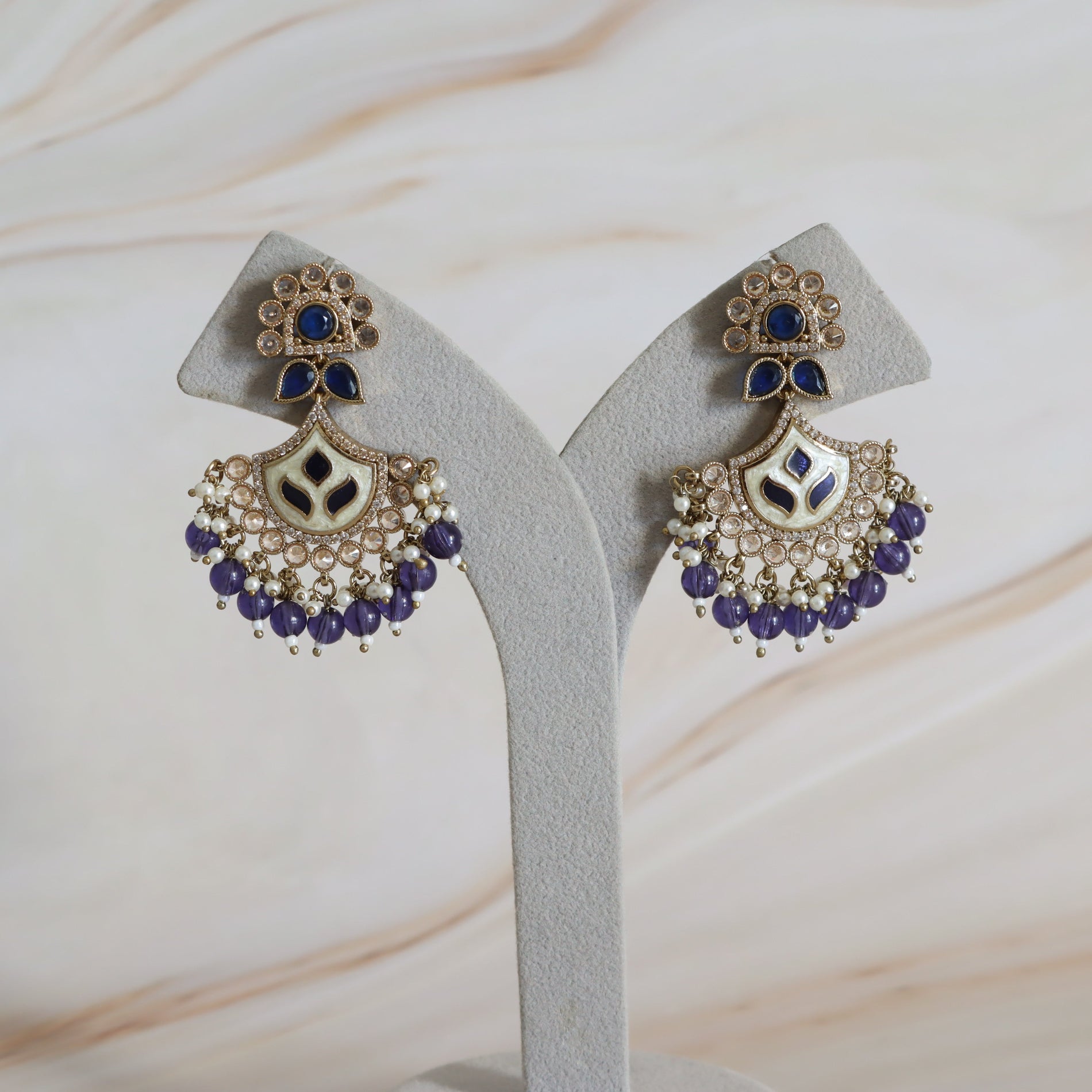 Pair of ornate earrings with blue and purple stones on a gray stand against a marble background.