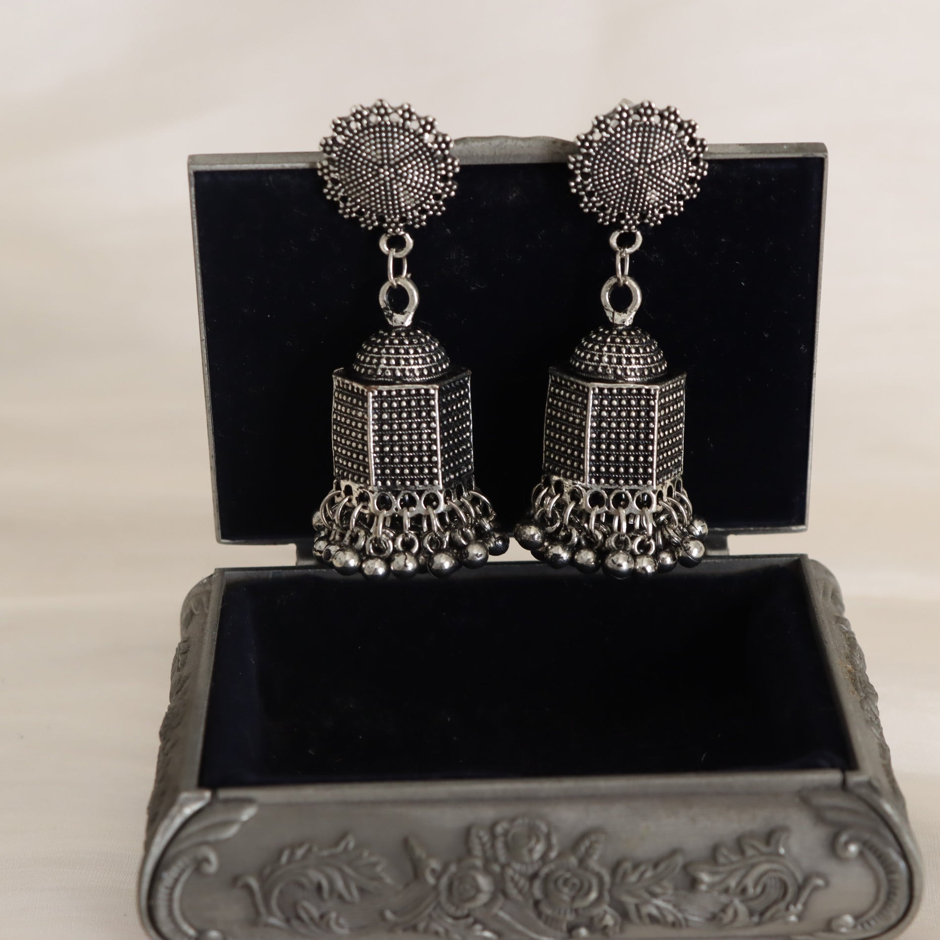 Pair of silver jhumka earrings on a decorative black box with floral patterns.