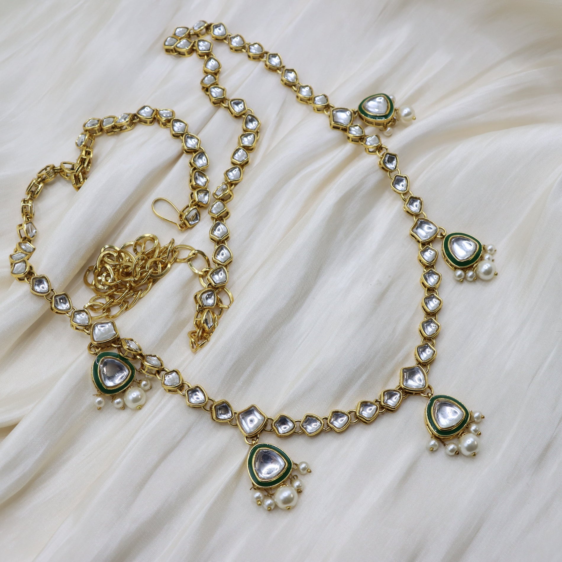 Waistband chain with clear teardrop-shaped stones, green enamel accents, and small white faux pearls, displayed on a cream-colored draped fabric background.